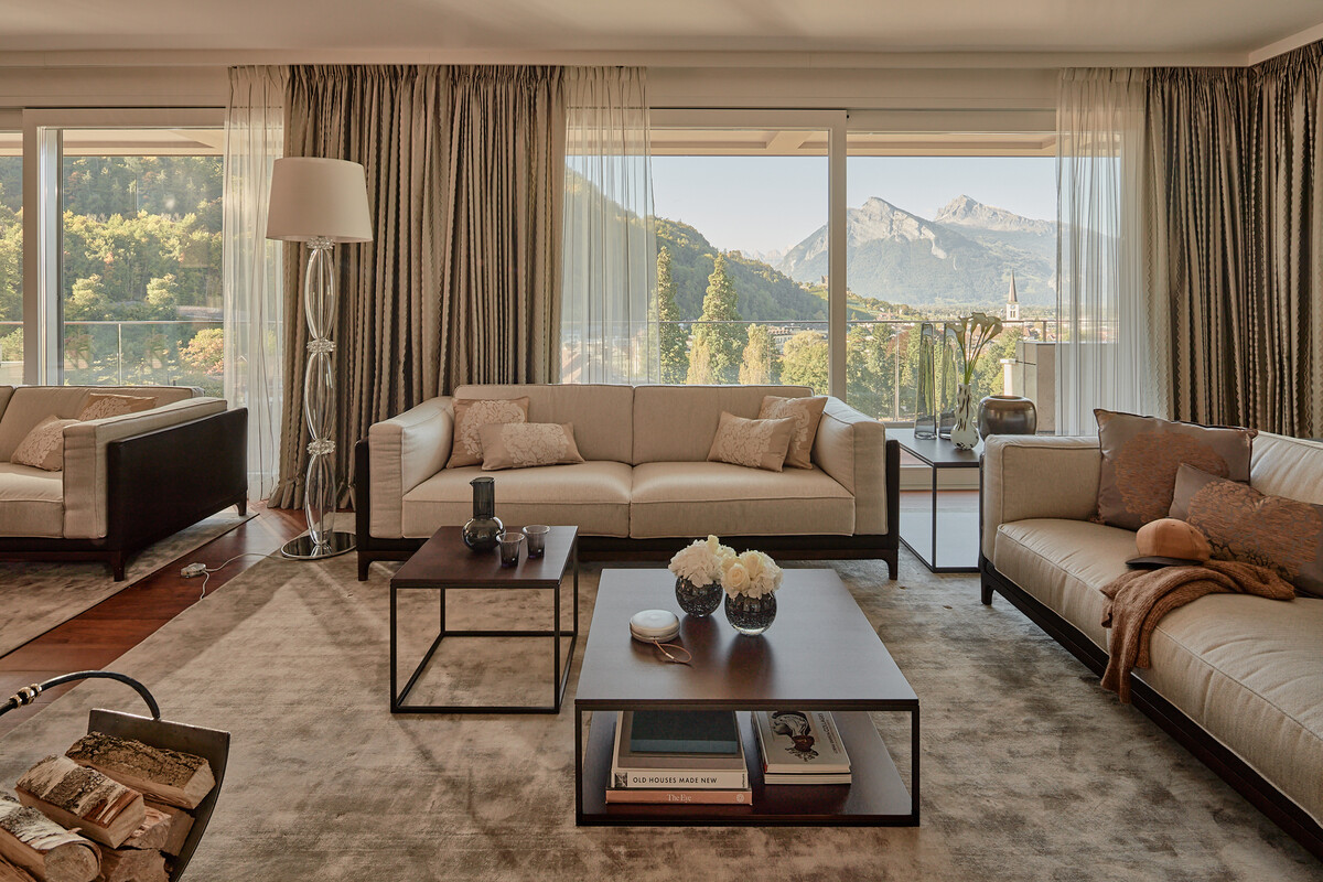 The morning sun's rays illuminate the living room of the Penthouse Suite in a reddish tone. Fresh flowers are on the living room table and mountains can be seen through the large windows. The Penthouse Suite is located at the top of the Spa Tower of the Hotel Grand Resort Bad Ragaz. This is a superior luxury resort in Switzerland.