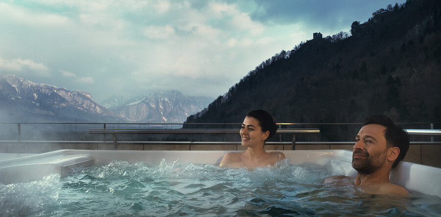Two guests sitting in an outdoor whirlpool at the parapet of a suite terrace, steam rising off the water with the valley and the ridge opposite in front of them.