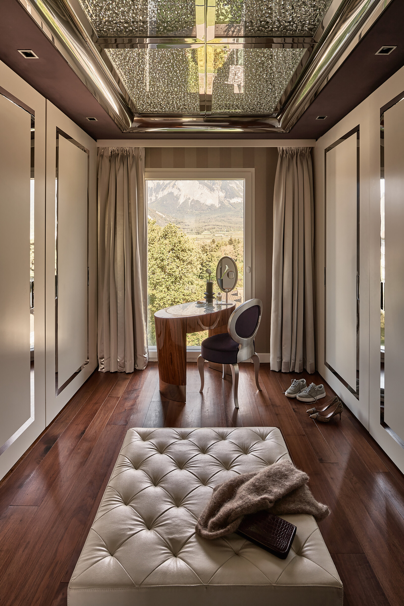 Luxuriöses Ankleidezimmer mit einer getufteten Bank im Vordergrund, einem schlichten Holztisch und einem verzierten Stuhl in der Nähe des Fensters. Der Blick auf die Berge, der Holzboden und die eleganten Vorhänge unterstreichen die ruhige Atmosphäre.