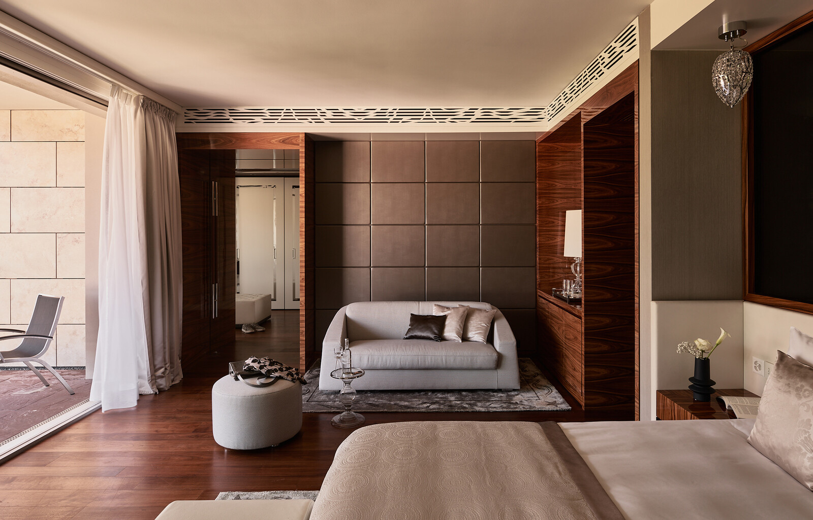 Luxuriöses Hotelzimmer in eleganten Braun- und Beigetönen. Mit modernem Bett, Glastisch, weissem Sofa und durchsichtigen Vorhängen, die auf einen Balkon führen. Gelassene Atmosphäre.