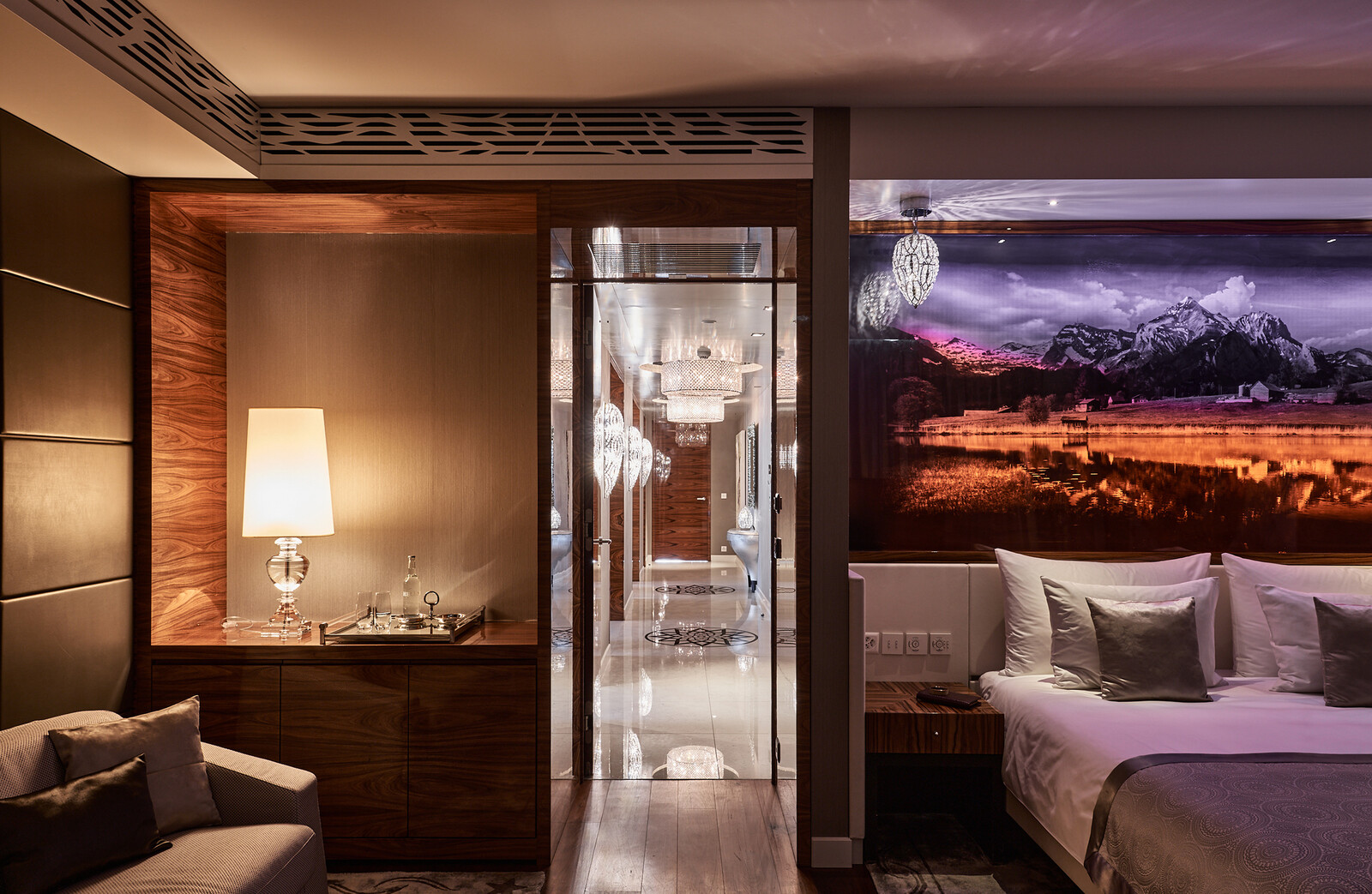 Luxuriöses Hotelzimmer mit warmem Ambiente, einem gemütlichen Bett mit weißen Kissen, einer beleuchteten Tischlampe auf einem Holzschrank und einer malerischen Berglandschaft als Wandbild.