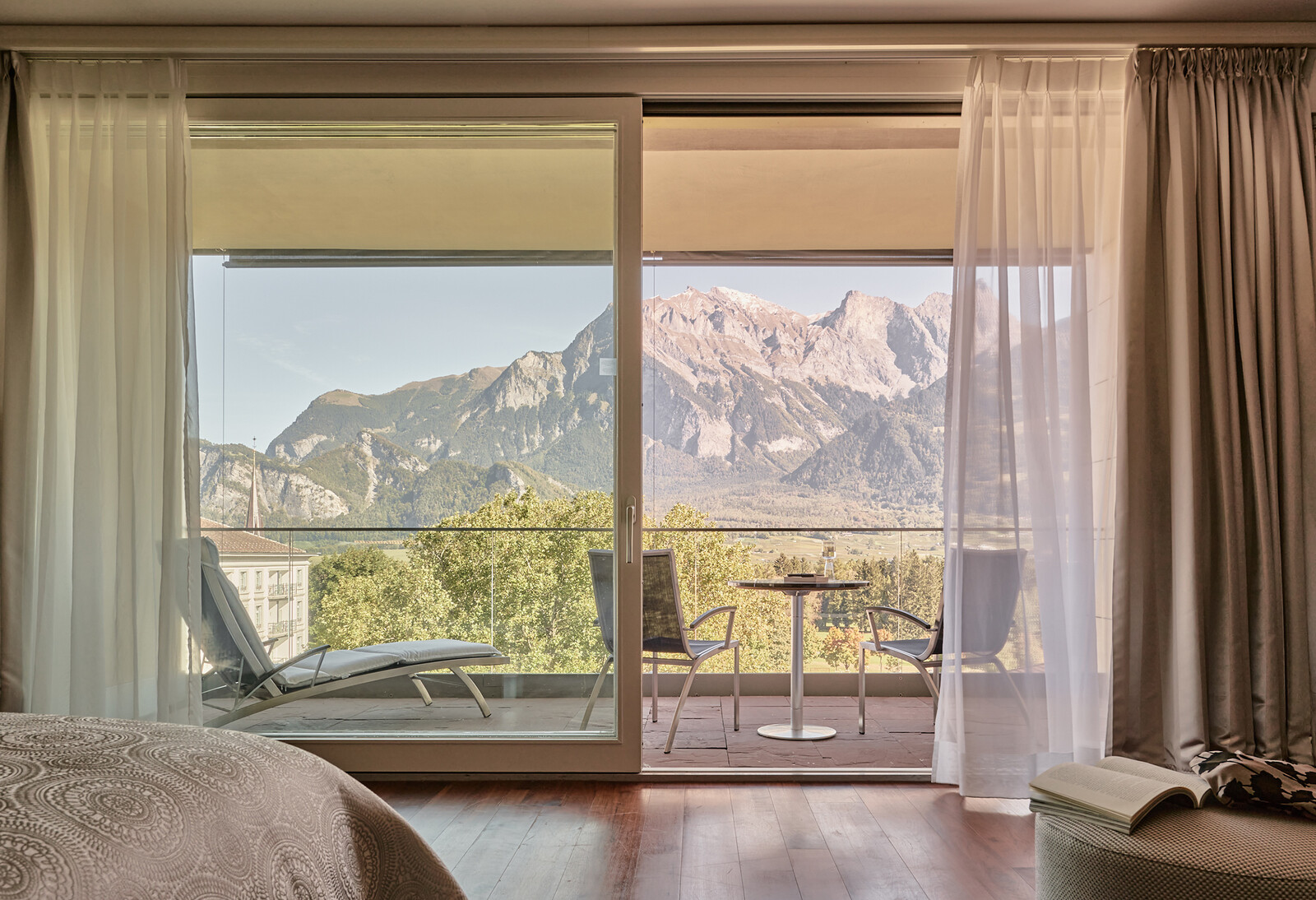Blick aus einem ruhigen Schlafzimmer mit einem Bett im Vordergrund auf einen Balkon mit zwei Stühlen, einem Tisch und einer herrlichen Berglandschaft hinter Glastüren.