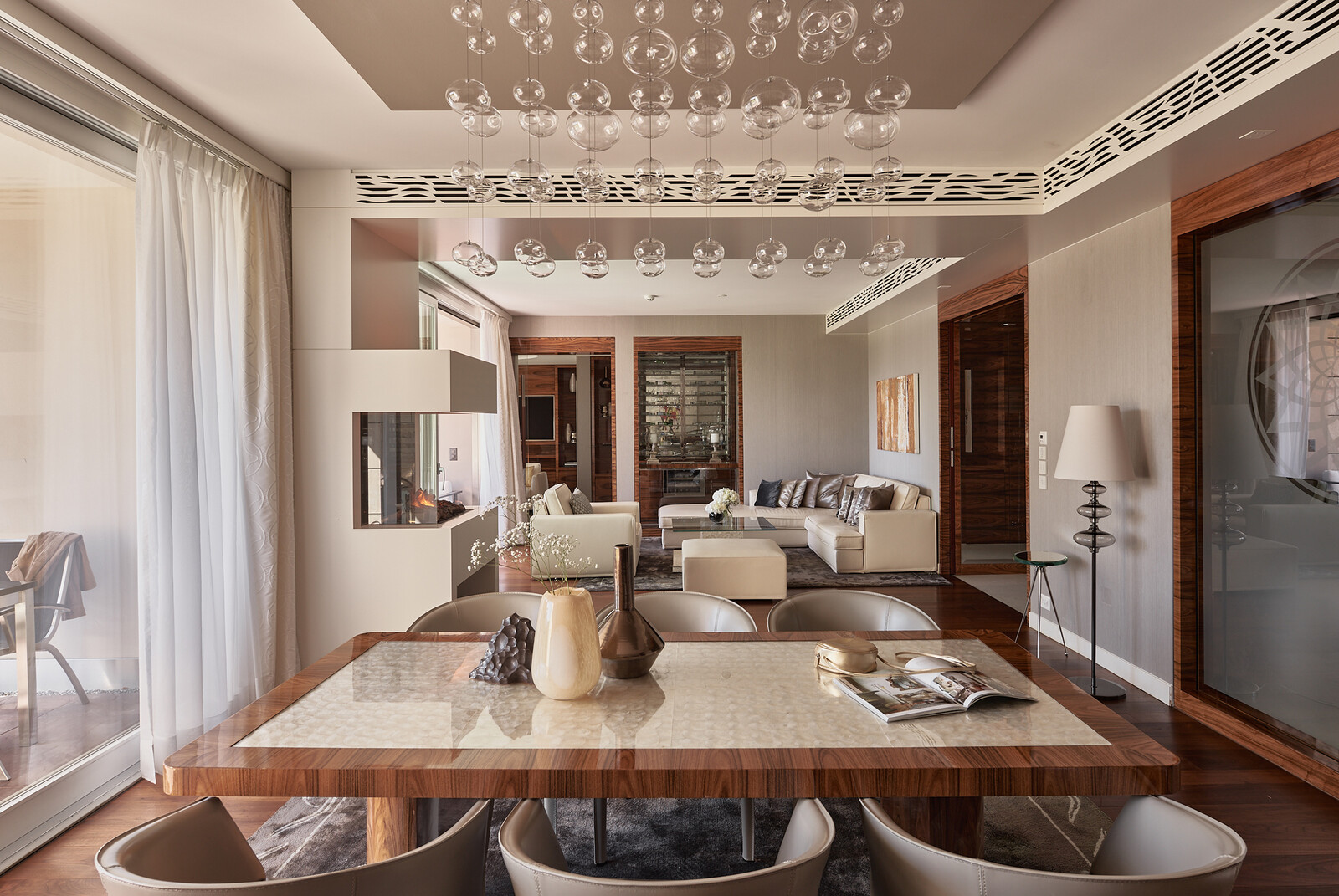 Modernes Wohnzimmer mit schickem Essbereich. Gläserne Pendelleuchten hängen über einem Holztisch mit Vasen. Im Hintergrund befindet sich eine gemütliche Sitzecke, die eine warme und einladende Atmosphäre schafft.