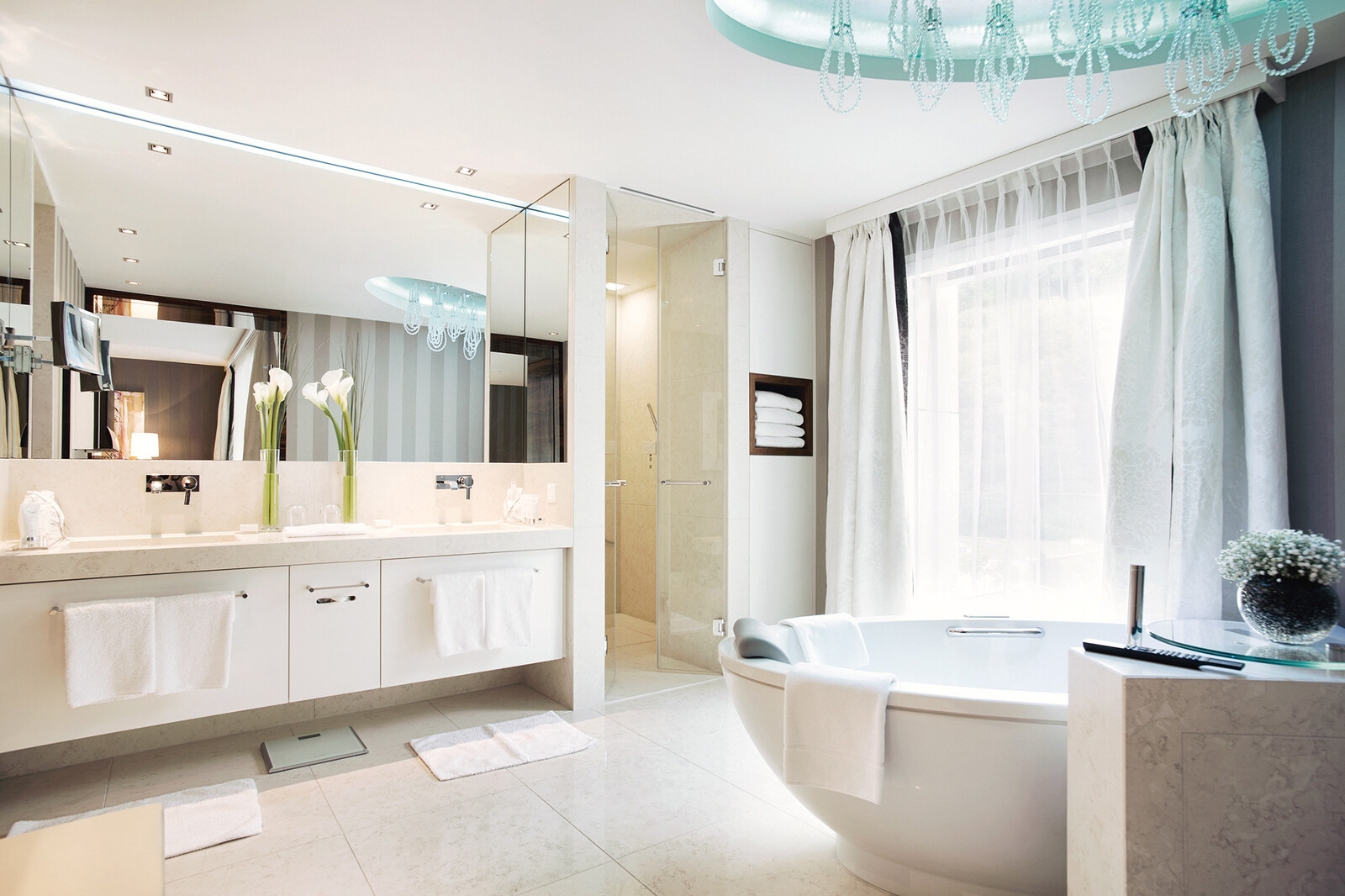 Luxuriöses, modernes Bad mit grosser runder Badewanne, elegantem Kronleuchter, Doppelwaschbecken und Glasdusche. Sanfte Beleuchtung und neutrale Töne schaffen ein ruhiges Ambiente.