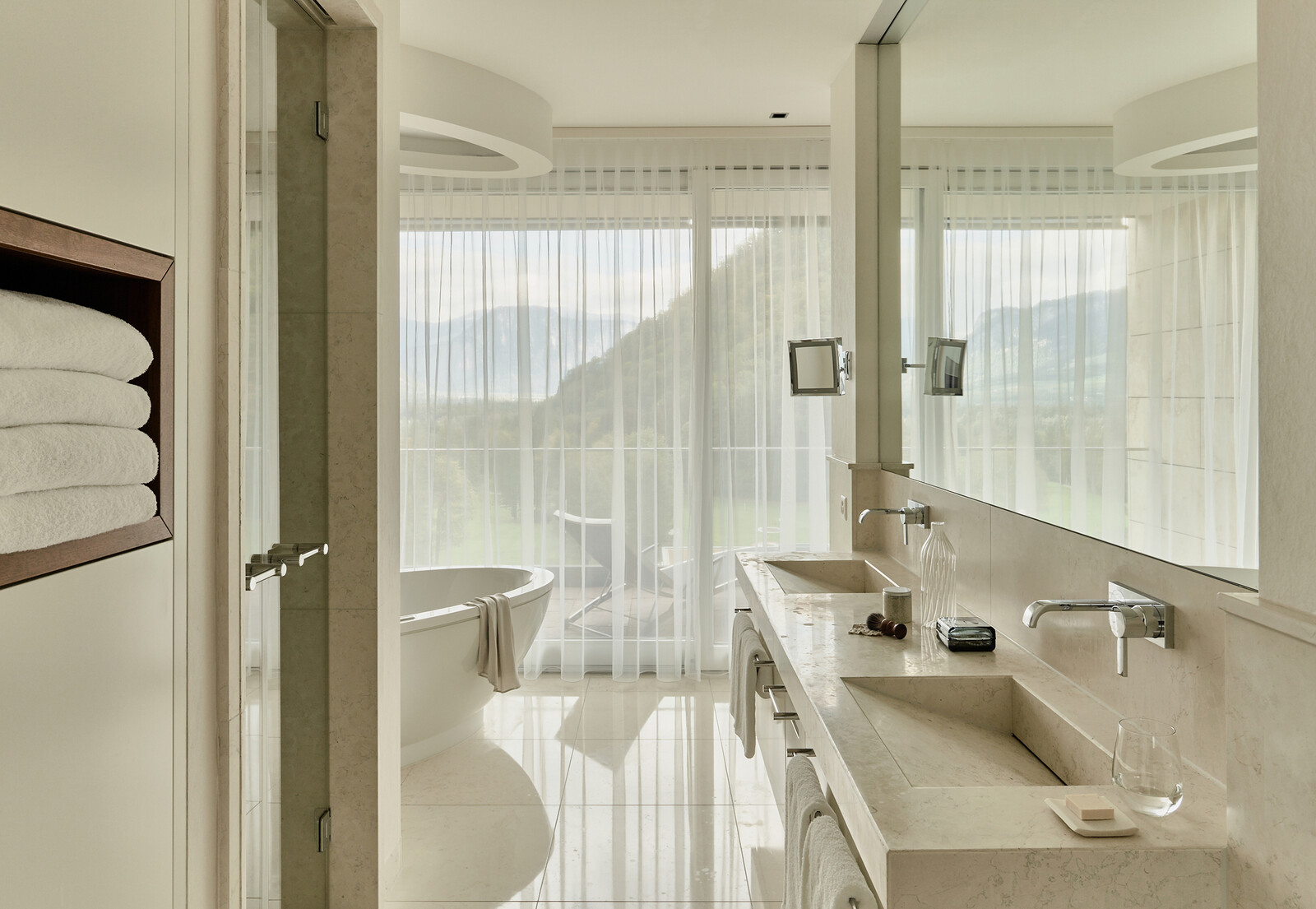 Ein modernes Badezimmer in hellen Farben mit zwei Waschbecken einer Badewanne und einem grossen Fenster direkt zum Balkon.