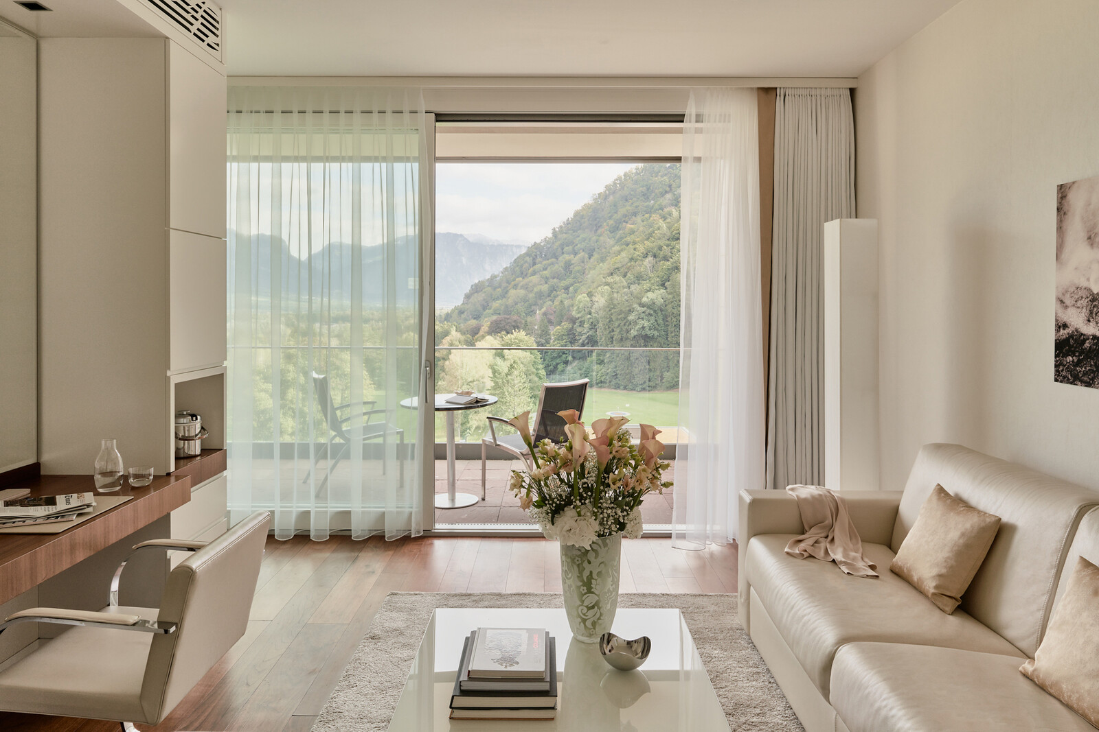 Modern living room with beige sofa, vase of flowers on a white coffee table, and sheer curtains framing a mountain view from sliding glass doors. Bright and serene.