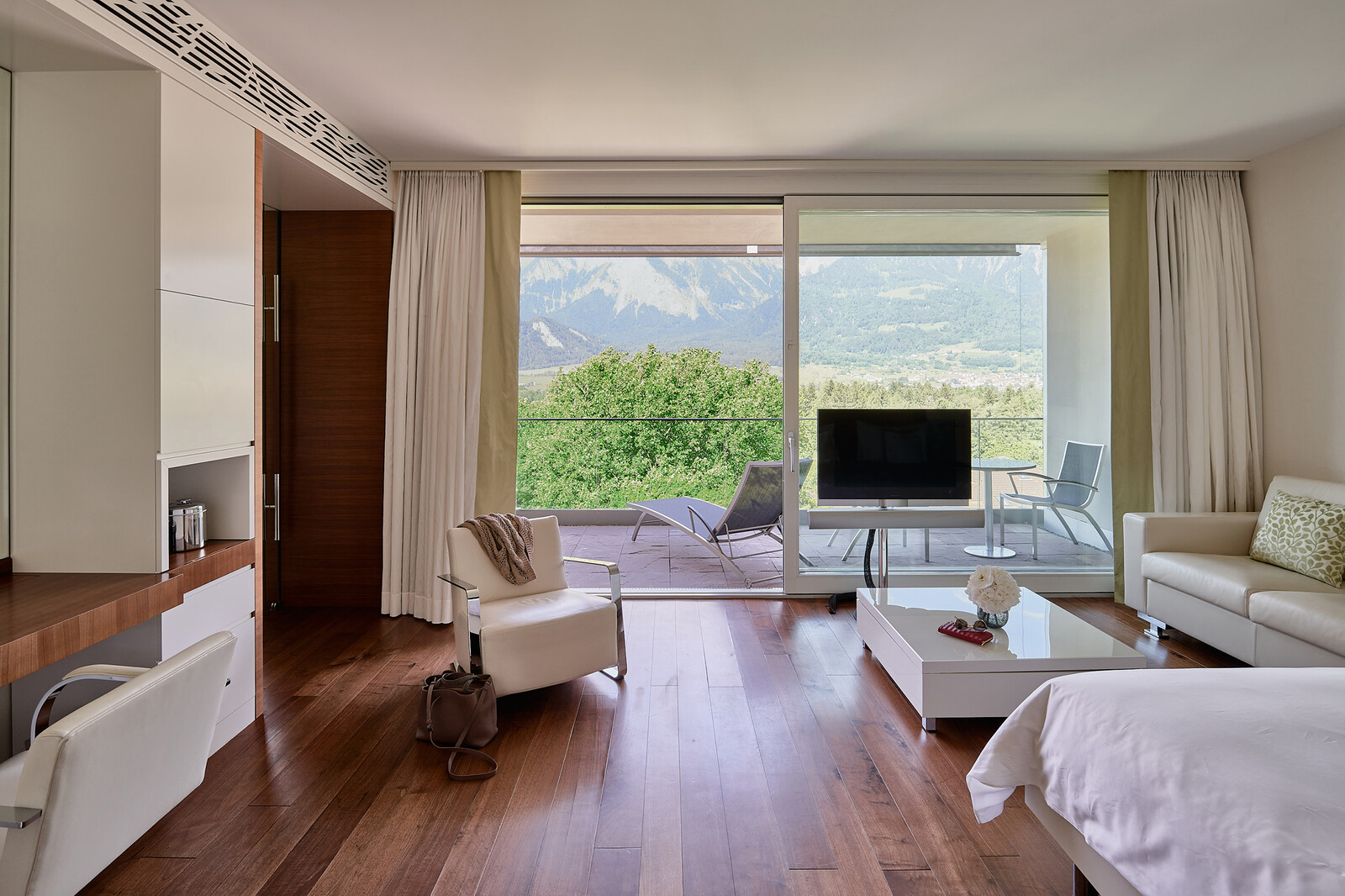 Modern hotel room with wooden floors, white furniture, and a glass wall revealing a balcony. The view includes lush greenery and mountains, creating a serene ambiance.