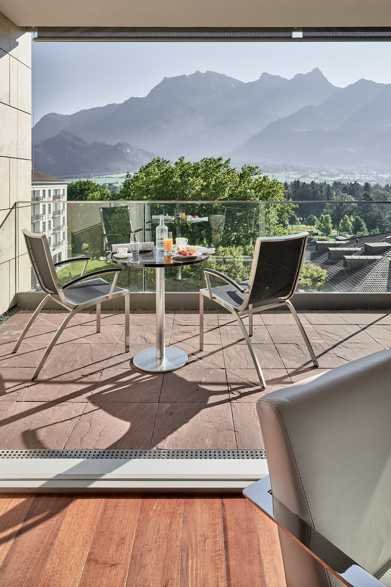 Balcony view of mountains with clear sky, two chairs and a table set for breakfast with juice and pastries, creating a serene and inviting atmosphere.