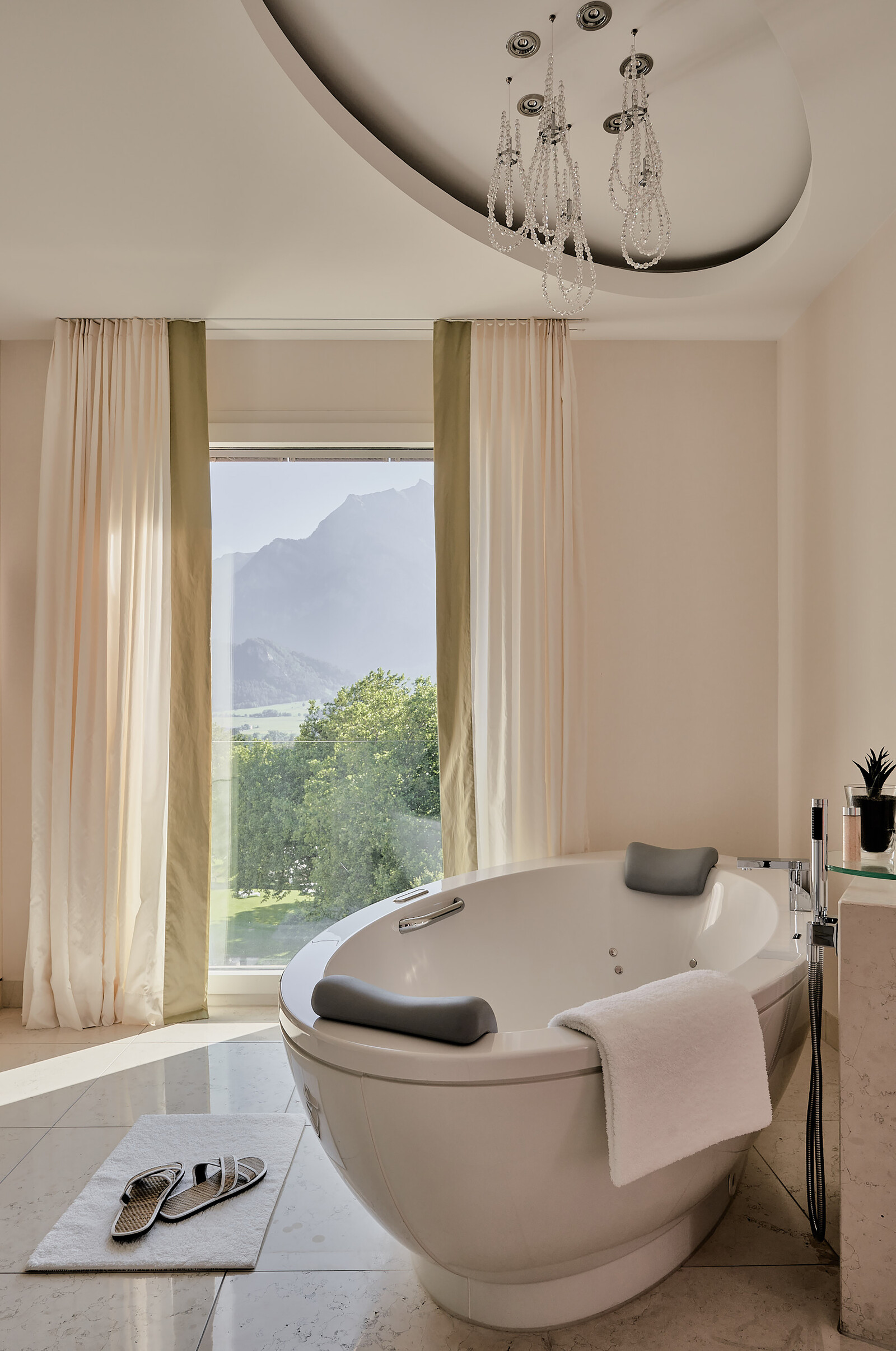 Luxurious bathroom with an oval bathtub beside a large window, offering a serene mountain view. Soft lighting and elegant chandelier create a tranquil ambience.