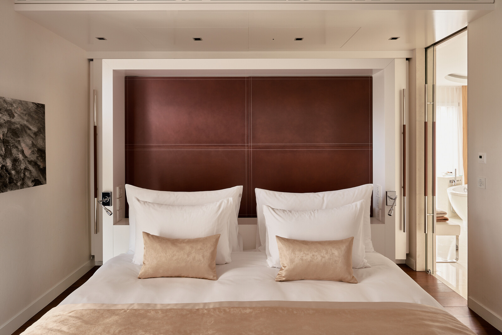 Luxurious hotel room with a large bed featuring crisp white linens and beige cushions. A brown padded headboard adds elegance, creating a serene, upscale ambiance.