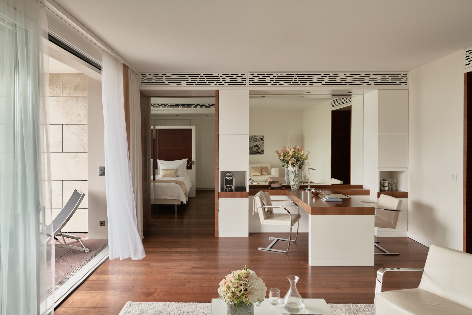 Modern, airy hotel suite with hardwood floors, open living space, elegant furniture, large windows with sheer curtains, and a bedroom in the background.