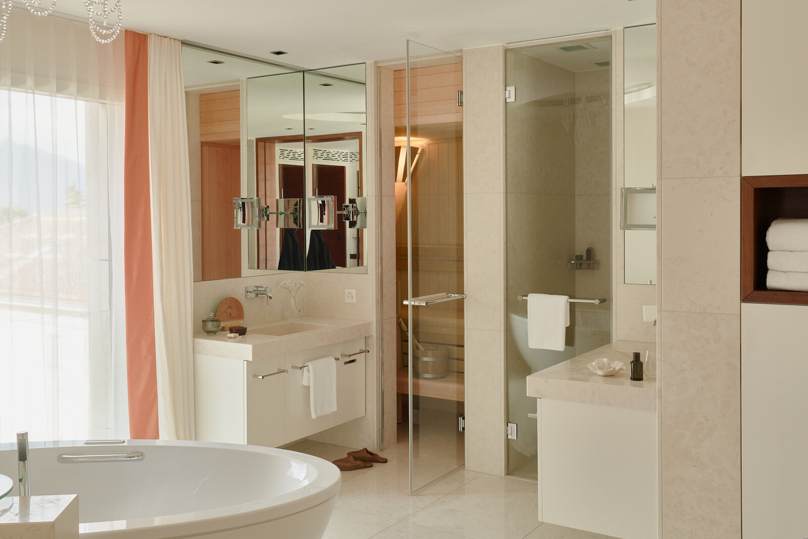 Modern bathroom with a glass shower, white sink, and large mirrors. A white bathtub is visible. The room has a calm, minimalist ambiance with soft natural light.