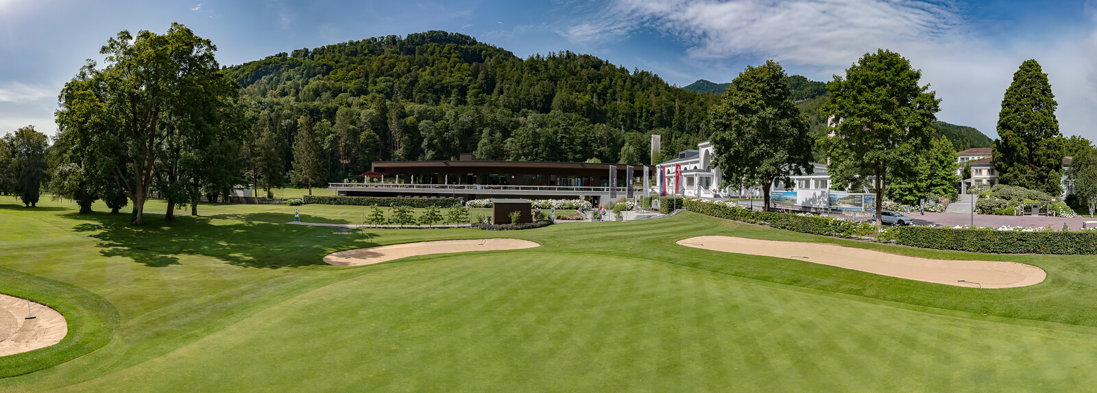 Panoramablick auf einen üppigen Golfplatz mit Sandbunkern im Vordergrund, umgeben von hohen Bäumen und einem Clubhaus. Im Hintergrund ist ein bewaldeter Hügel unter einem teilweise bewölkten Himmel zu sehen.