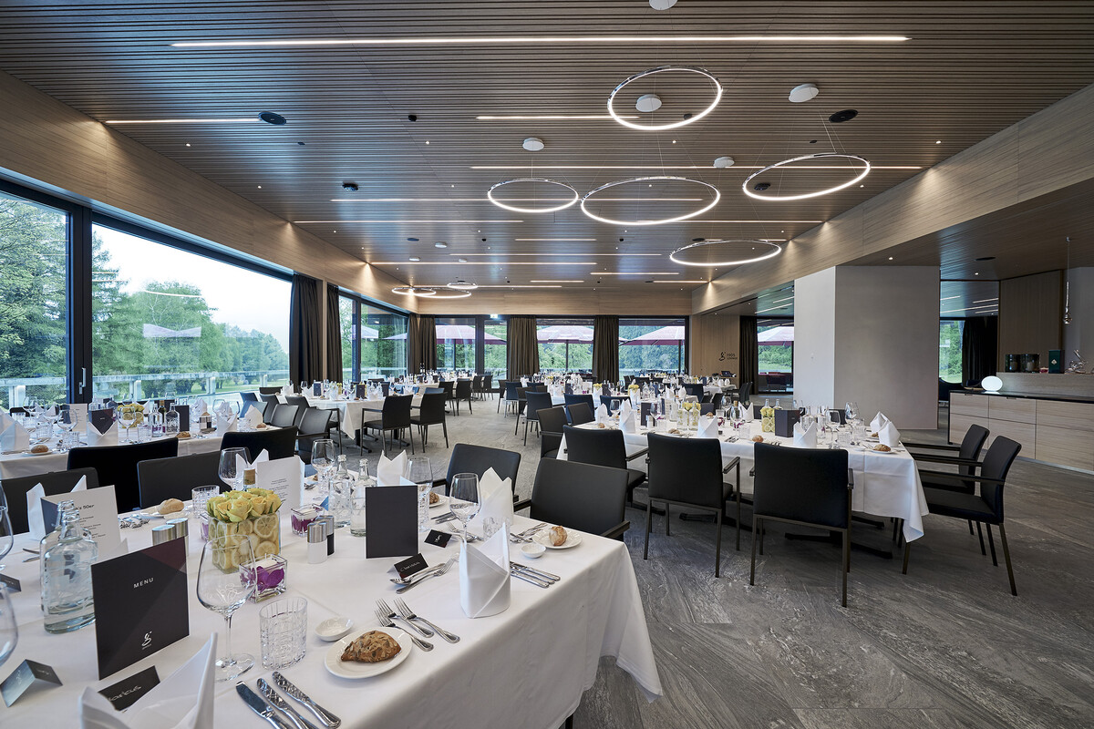 Ein geräumiger Restaurantsaal mit elegant gedeckten Tischen mit weißer Bettwäsche, schwarzen Stühlen und modernen, kreisförmigen Leuchten bietet ein ruhiges, gehobenes Ambiente.