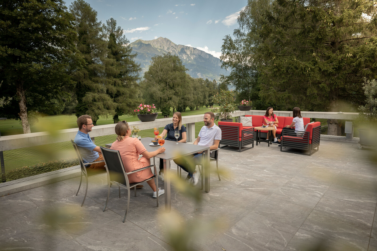 Aussenterrasse mit sechs Personen, die sich entspannen und sich unterhalten. Zwei Gruppen sitzen an Tischen, umgeben von hohen Bäumen und Bergen, die eine ruhige Atmosphäre schaffen.