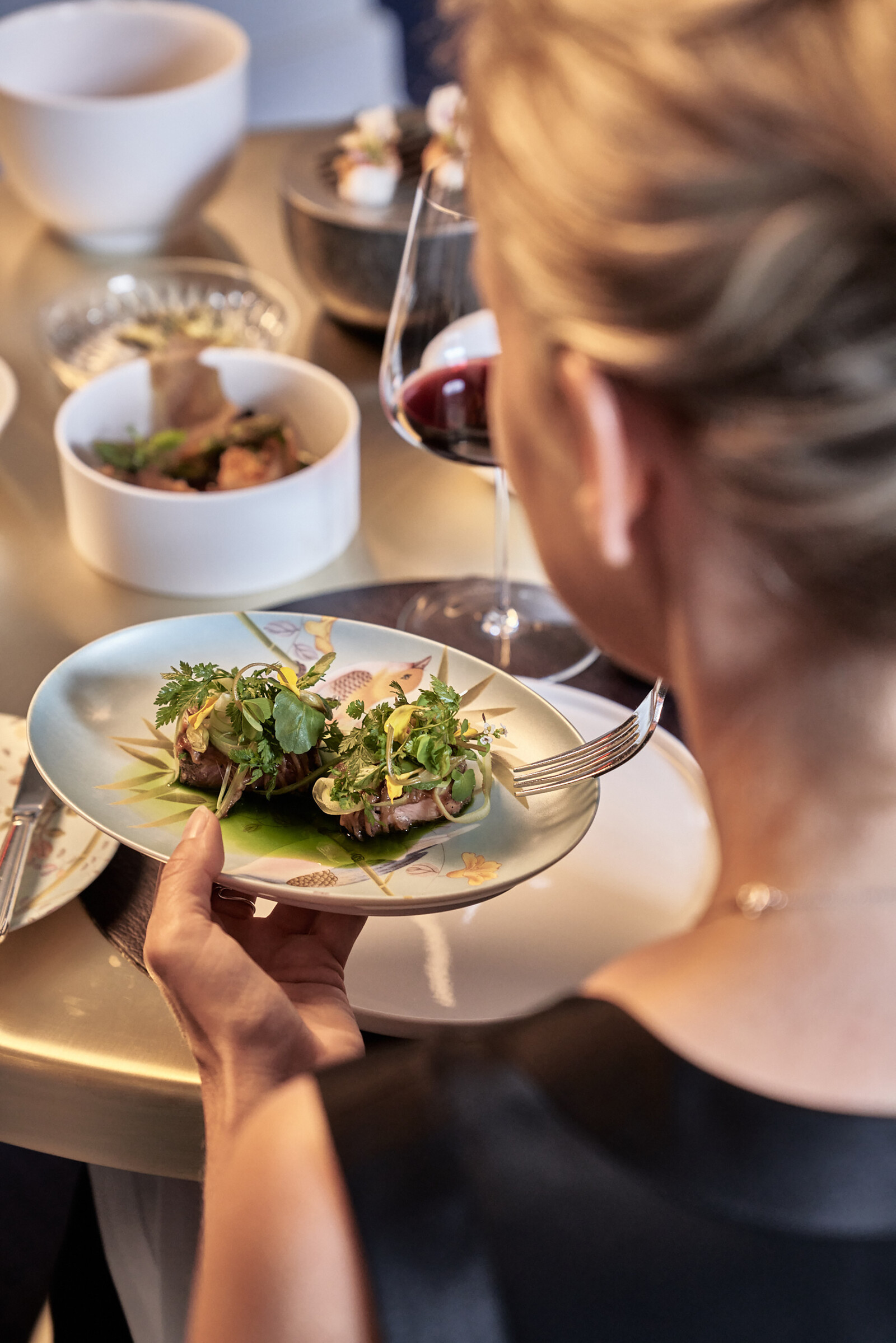 Das Gericht wurde mit Blättern und Kräutern serviert. Der Gast, eine blonde Dame, ist im Begriff dieses mit ihrer Gabel zu verspeisen. Das Restaurant Igniv by Andreas Caminada ist eines der Restaurants des Hotel Grand Resort Bad Ragaz, einem Superior Luxusresort in der Schweiz.