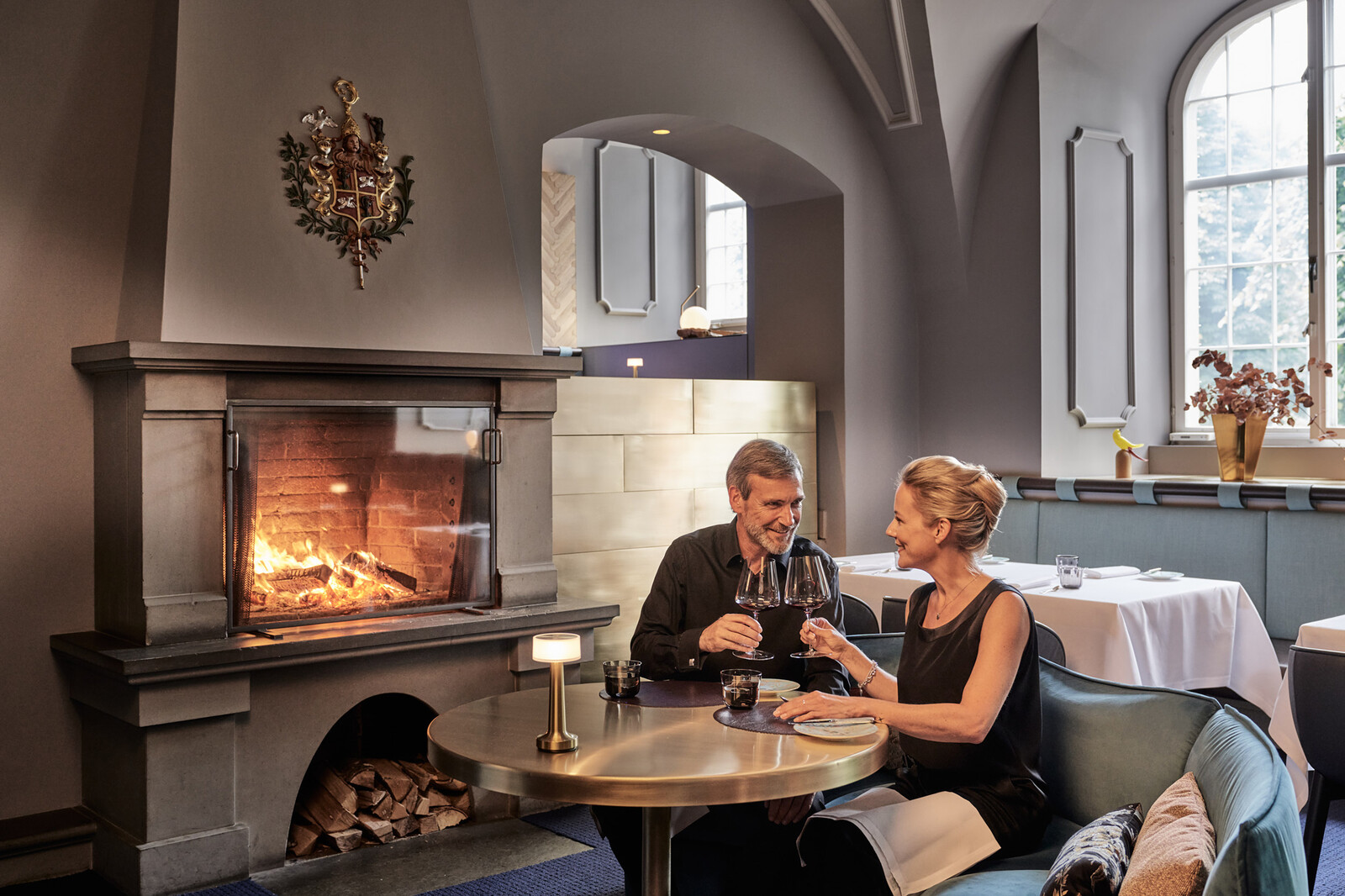 Der kleine runde Tisch befindet sich neben einem Kamin, in dem ein Feuer brutzelt. Zwei Gäste stossen mit Weingläsern an. Das Restaurant Igniv by Andreas Caminada befindet sich im Hotel Grand Resort Bad Ragaz, einem Superior Luxusresort in der Schweiz.
