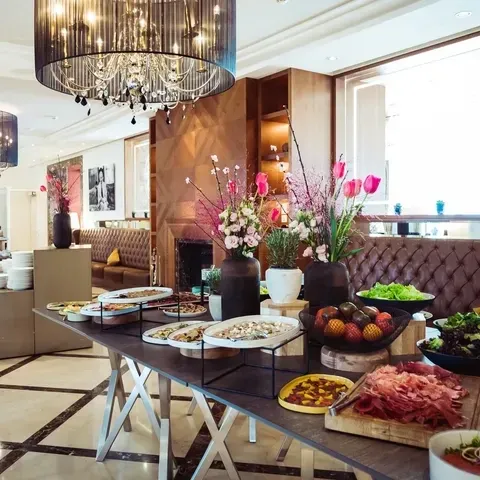 Luxuriöses Buffet mit Tellern voller verschiedener Speisen auf einem langen Tisch, eleganten Kronleuchtern an der Decke und Vasen mit bunten Blumen, die einen Hauch von Raffinesse verleihen.