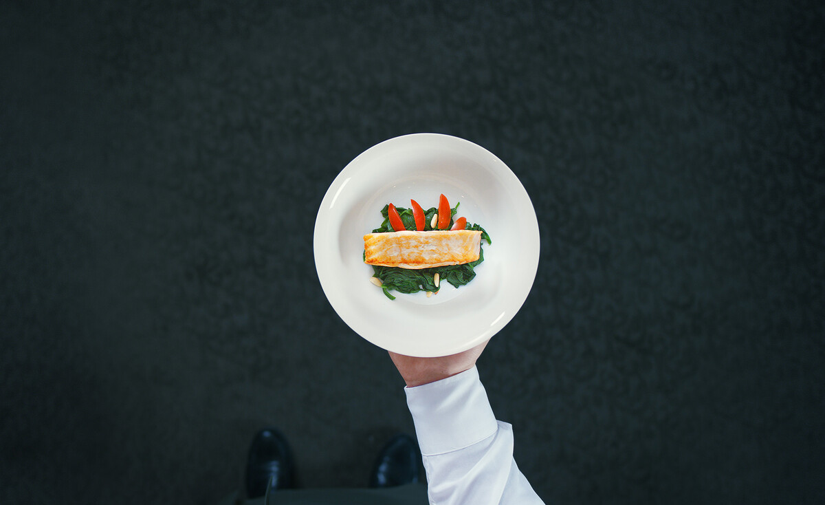 Ein Koch präsentiert Fischgericht mit Lachs und Spinat auf einem weißen Teller.