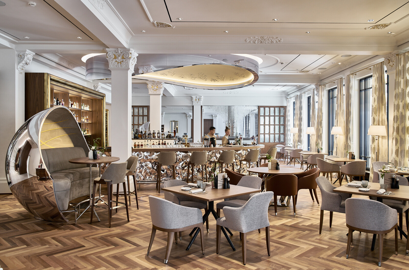 Elegantes Restaurant mit modernem Dekor, einer zentralen Bar, modernen Sitzgelegenheiten, großen Fenstern und warmer Beleuchtung, die eine einladende Atmosphäre schaffen.