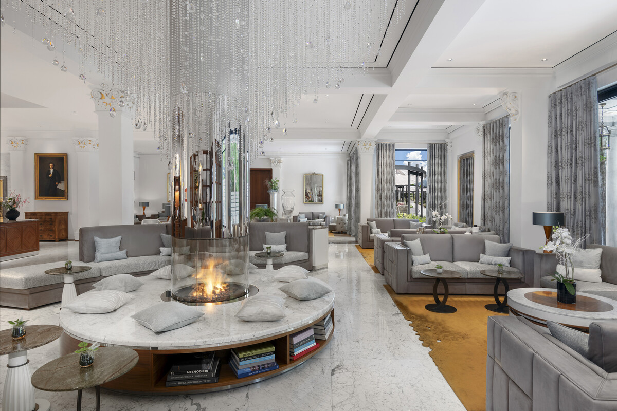 Luxuriöse Lobby des Quellenhof Bad Ragaz mit einem zentralen runden Kamin, umgeben von grauen Sofas und Kissen. Darüber hängen Kristallkronleuchter, die ein elegantes Ambiente schaffen.