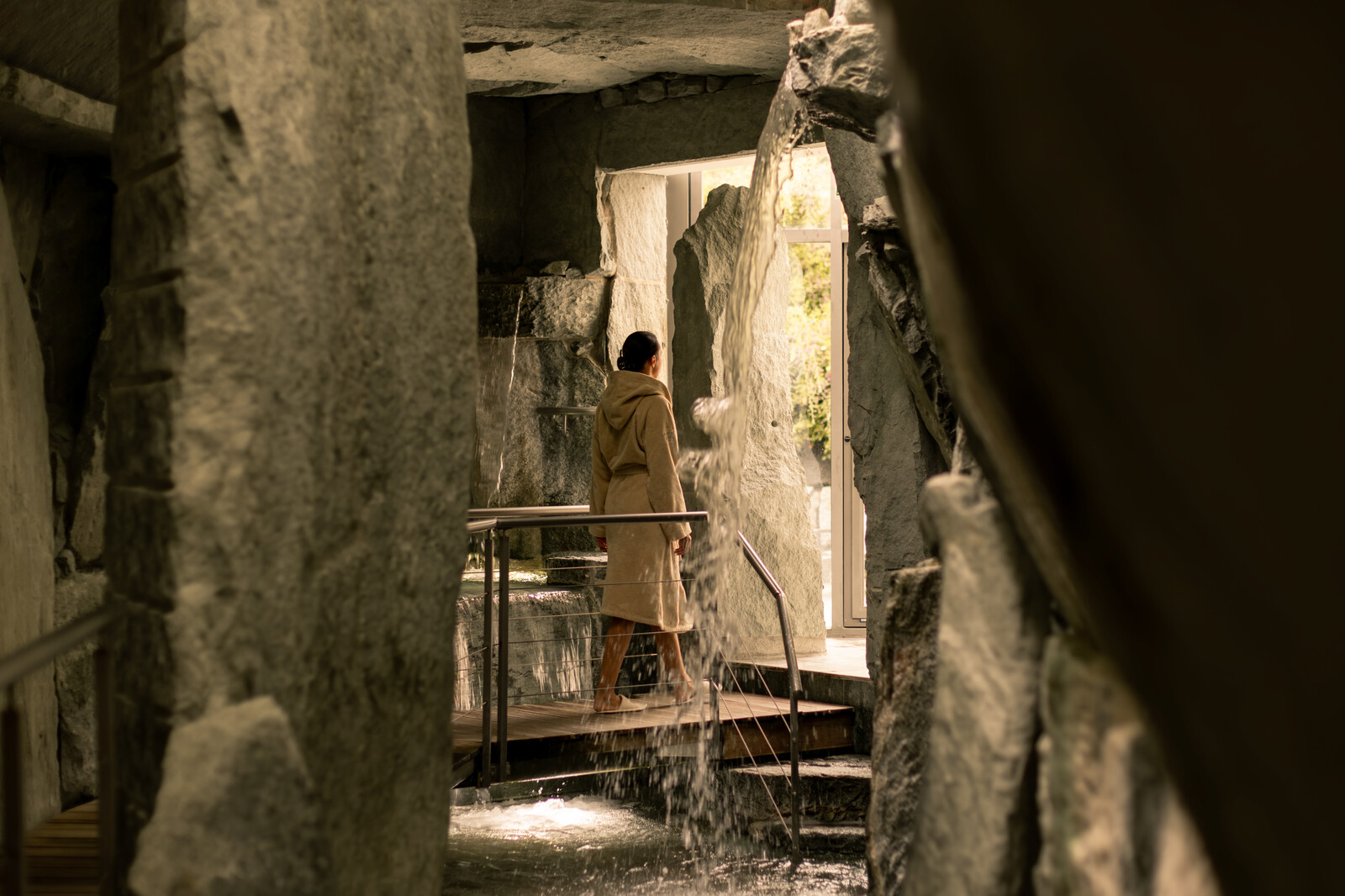 Person in a robe stands on a narrow wooden path surrounded by rocky walls and cascading waterfalls, gazing at sunlight streaming through a doorway. Serene and contemplative.