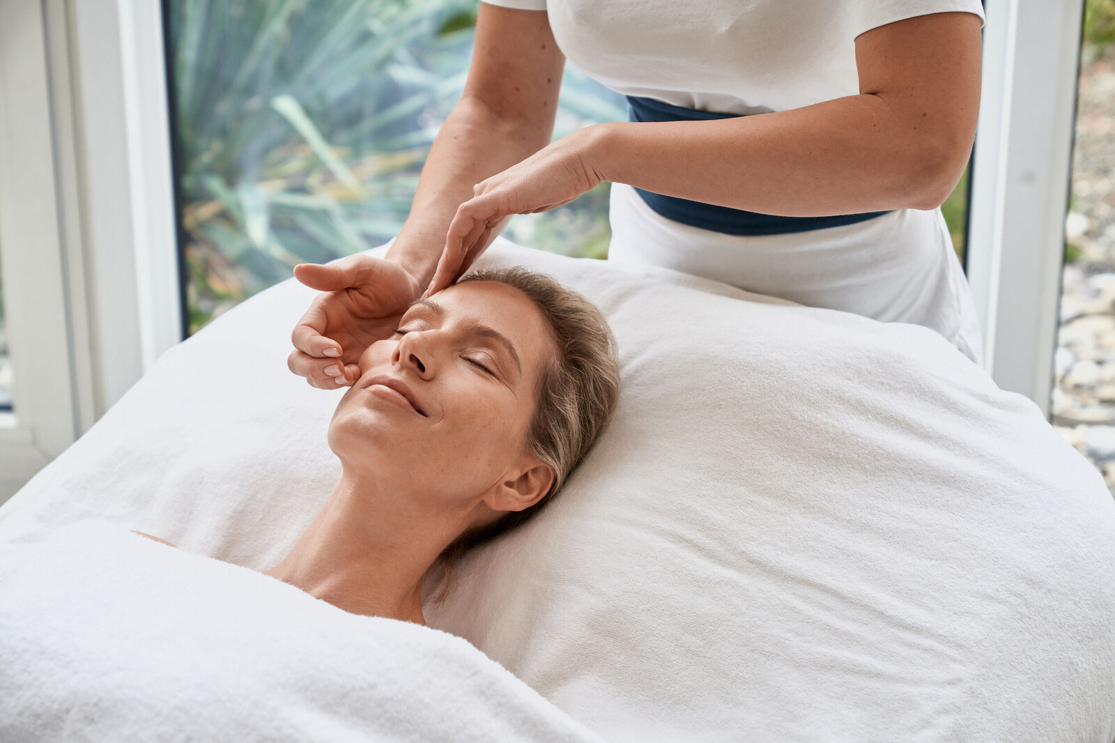 Die Kosmetikerin gibt dem Gast, einer Frau, ein Facial Retreat, während diese auf dem Rücken liegt, bedeckt mit Tüchern. Der Behandlungsraum, in dem sich die beiden befinden, gehört zum Thermal Spa, dem Spa des Hotel Grand Resort Bad Ragaz, welches ein Superior Luxusresort in der Schweiz ist.