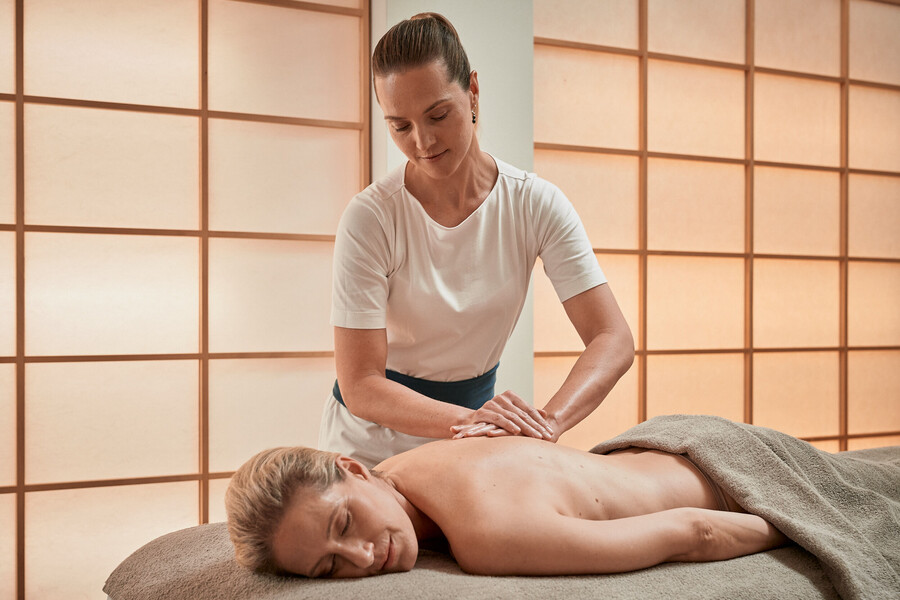 A therapist working with both hands flat on a guest's back, who lies face down under a grey towel on a treatment couch in front of a translucent screen wall.