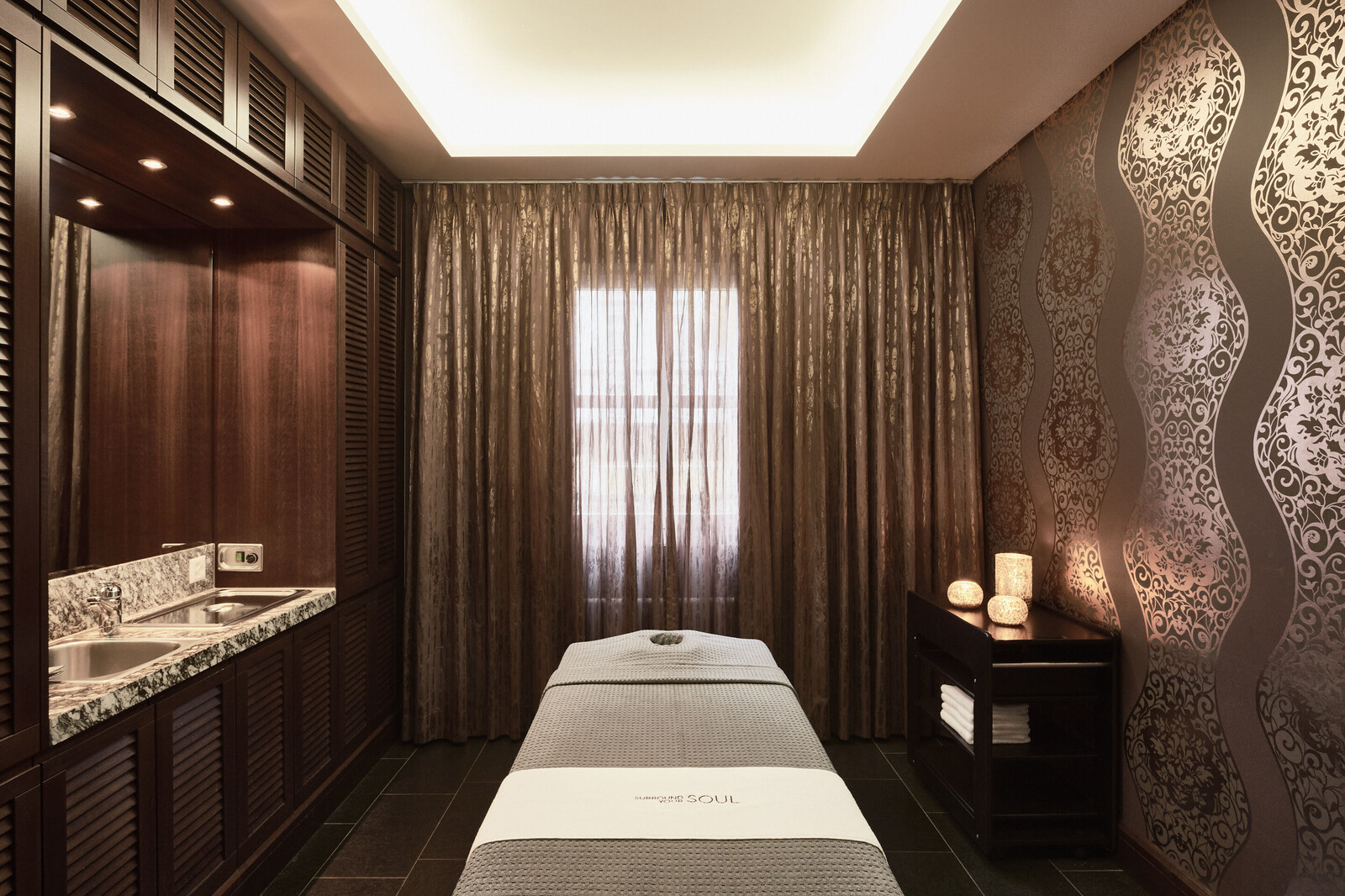 Der Behandlungsraum Pacific ist von vorne zu sehen. Er ist in Braun gehalten und dezenter goldener Verzierung. Eine Liege steht in der Mitte des Raumes, welcher sich im Thermal Spa des Hotel Grand Resort Bad Ragaz, einem Superior Luxusresort in der Schweiz, befindet.