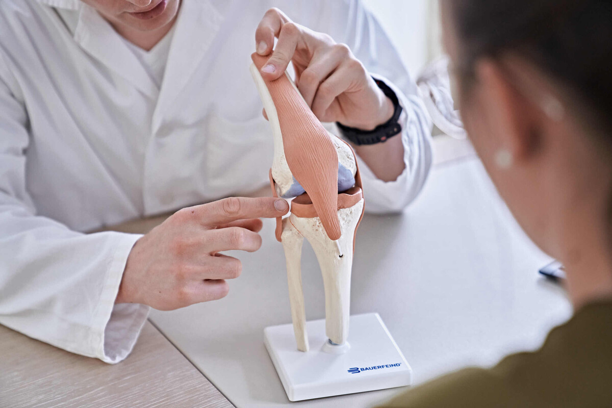 Blick über die Schulter einer Patientin auf ein künstliches Gelenk. Ein Arzt zeigt mit den Fingern darauf und erklärt etwas.