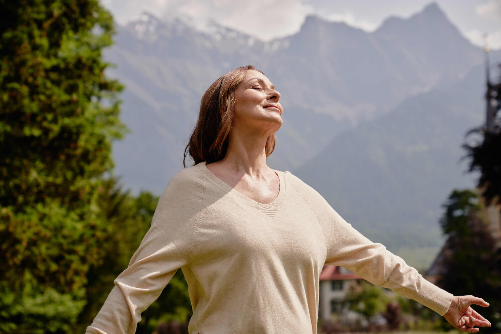 Blick auf eine Frau mittleren Alters, die zufrieden ihre Hände über ihr Herz legt. Im Hintergrund ist eine Bergkulisse zu sehen.