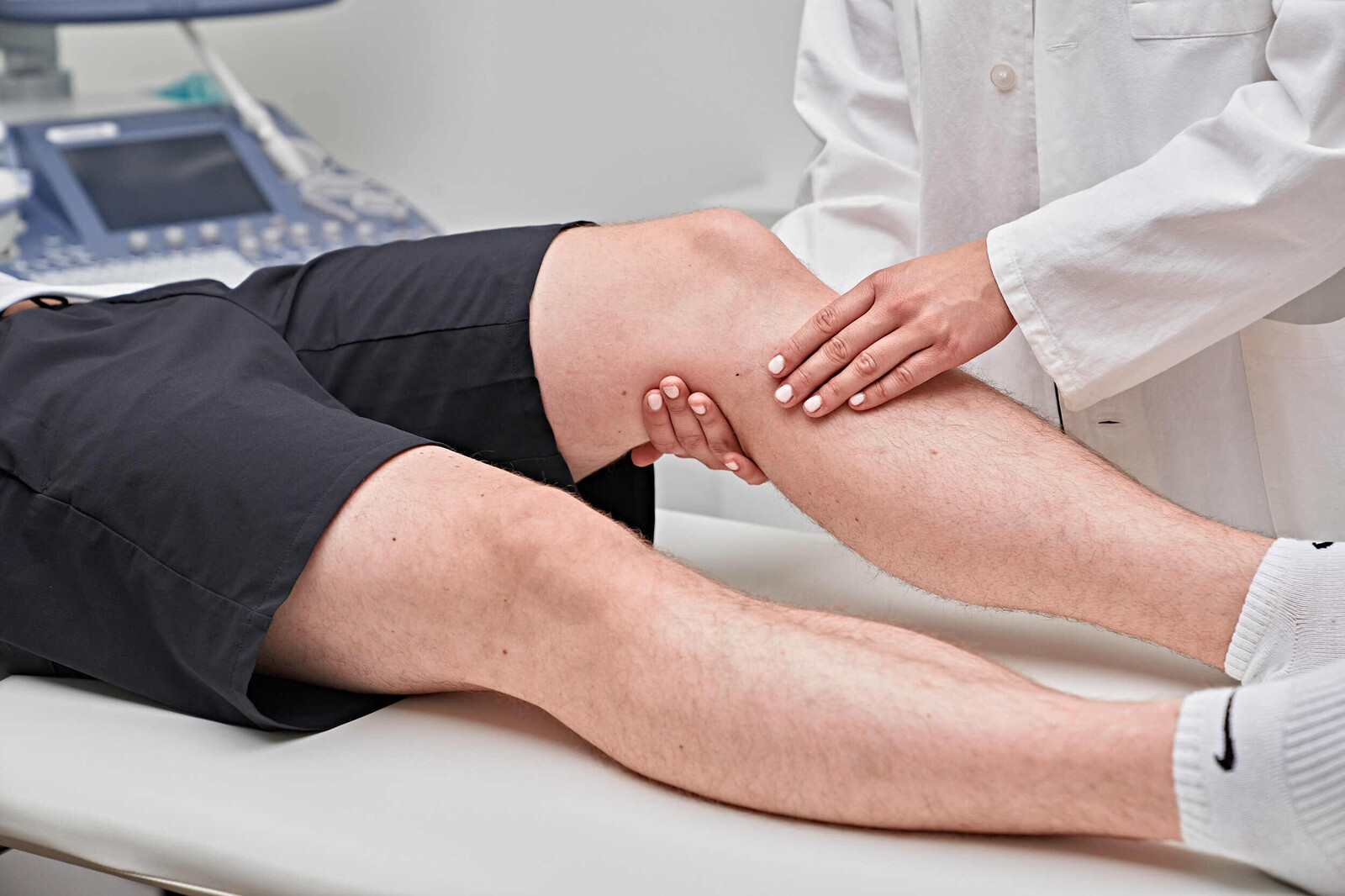 Blick auf die Beine eines liegenden Patienten. Eine Ärztin tastet dessen Knie ab.