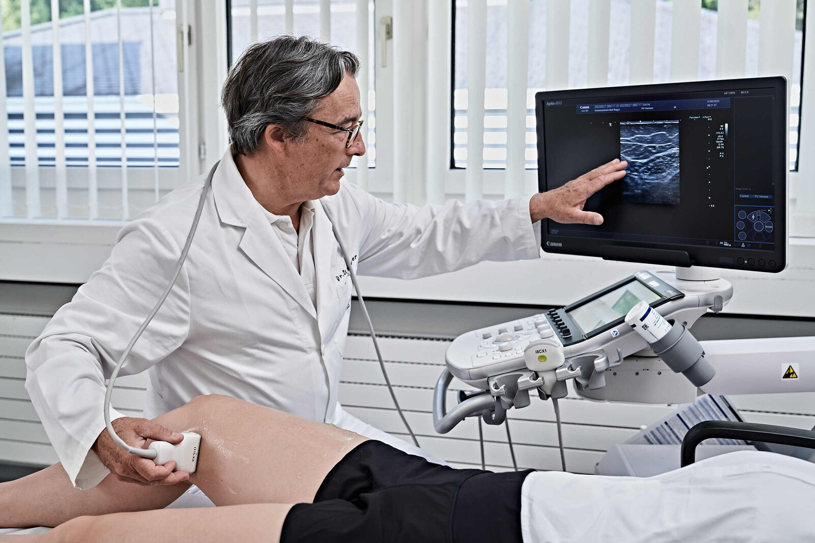 Ein Arzt untersucht mittels Ultraschall das Knie eines Patienten, der auf einer Liege liegt. Der Arzt erklärt, was er sieht anhand eines Bildschirms, der den Ultraschall zeigt.