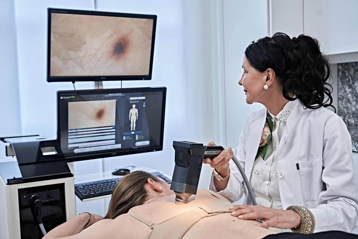 Eine Patientin liegt auf ihrem Bauch, während ihre Muttermale von einer Hautärztin untersucht werden. Auf den Bildschirmen im Hintergrund werden die Muttermale vergrössert dargestellt.
