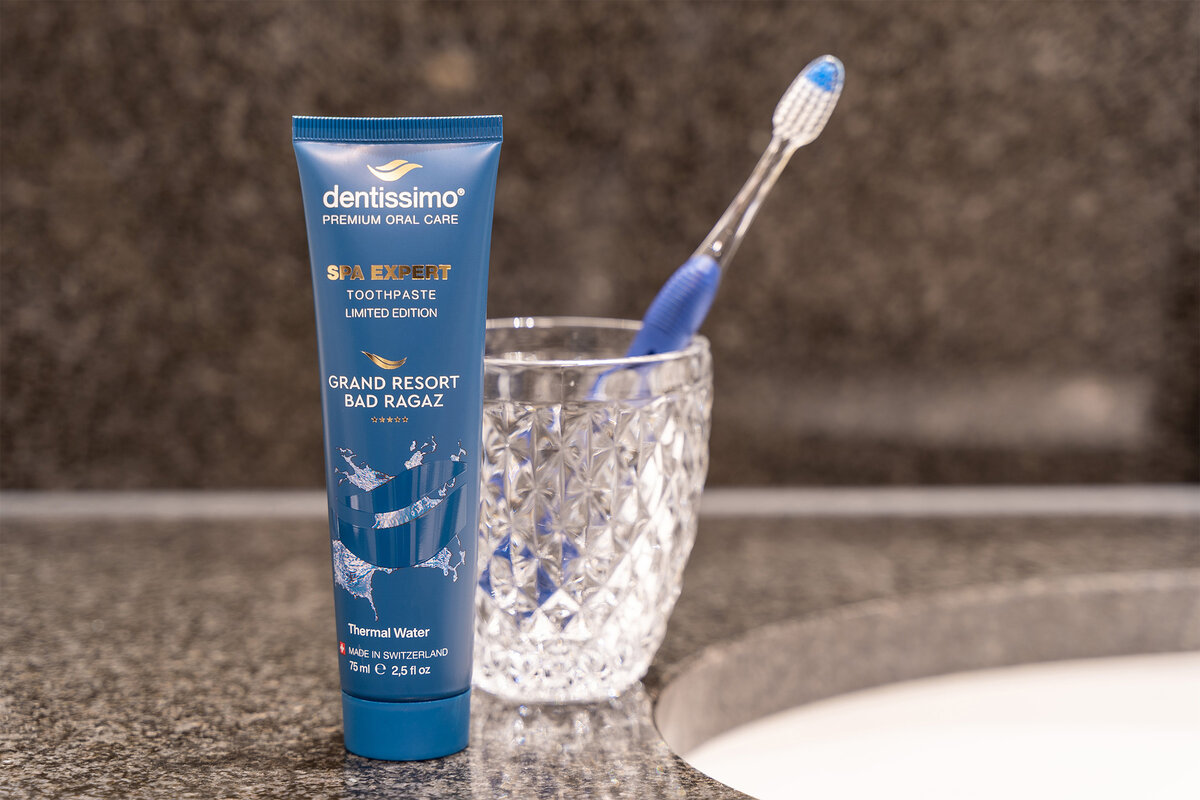 Eine blaue Zahnpastatube mit der Aufschrift „Dentissimo Spa Expert” steht auf einer Granitarbeitsplatte neben einem Glas mit Diamantmuster, in dem eine blaue Zahnbürste steckt. Sauber und elegant.