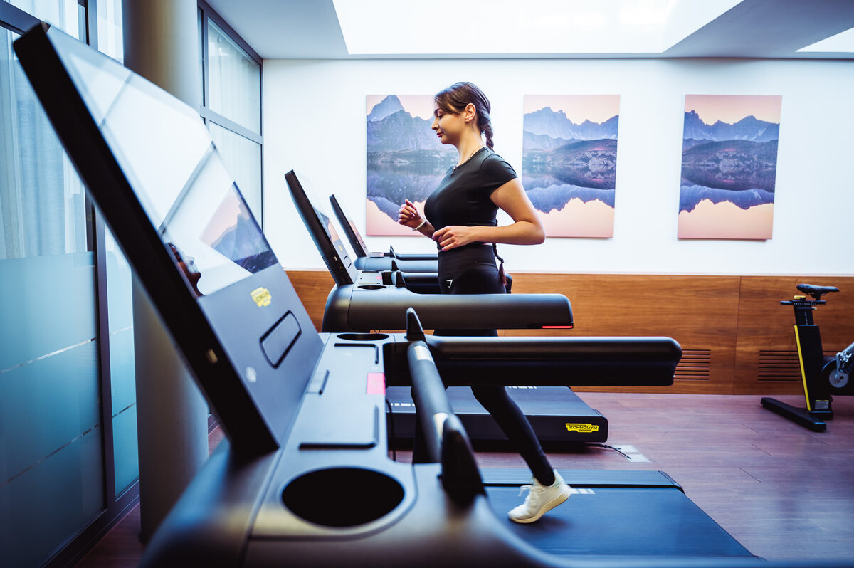Eine Frau joggt auf einem Laufband in einem hellen, modernen Fitnessstudio. Große Landschaftsfotos schmücken die Wand.