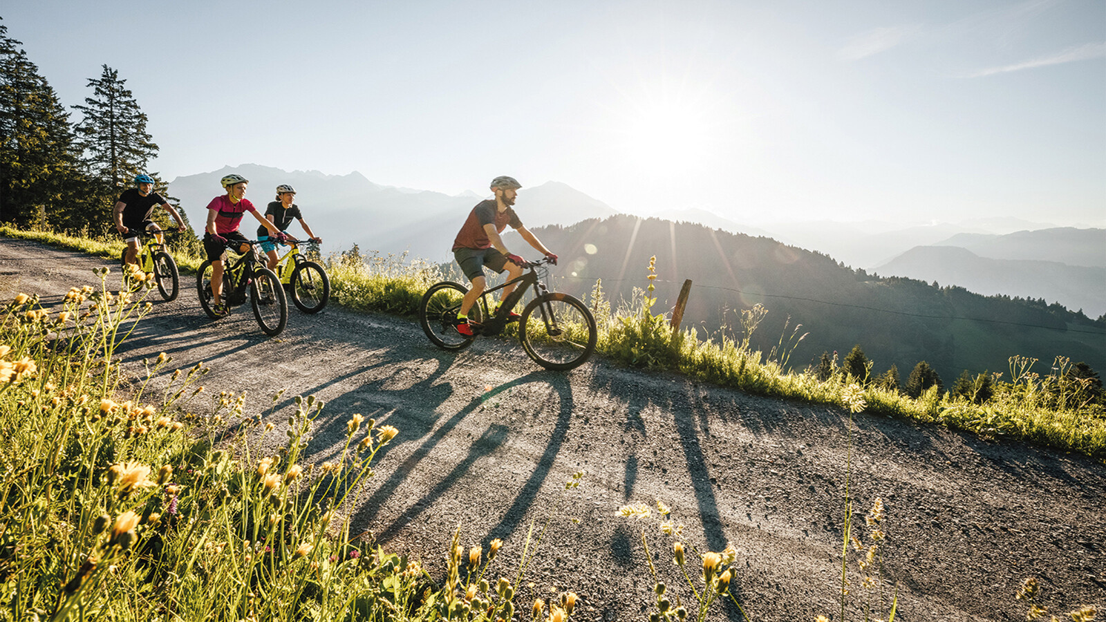 Vier Mountainbiker fahren einen Weg hinunter. Die Sonne geht bald unter, steht aber noch am Himmel. Das Gras am Wegrand ist grün. Bäume und Berge umgeben die Biker. Diese Umgebung befindet sich in der Nähe des Hotel Grand Resort Bad Ragaz, einem Superior Luxusresort in der Schweiz.