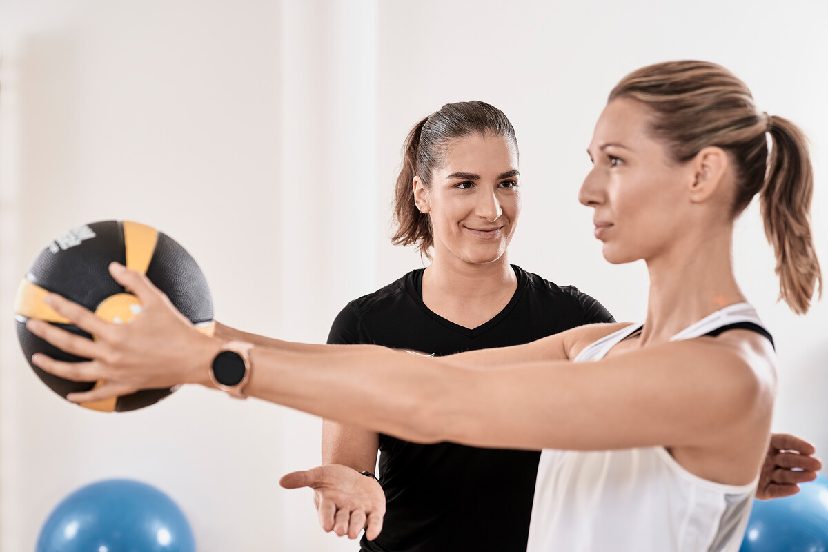 Ein Gast hält einen Ball mit gestreckten Armen vor sich. Die Therapeutin unterstützt sie dabei, indem sie ihre Körperhaltung korrigiert. Mit Hintergrund liegen blaue Gymnastikbälle auf dem Boden. Das Fitnesscenter befindet sich im Grand Resort Bad Ragaz, einem Superior Luxushotel in der Schweiz.