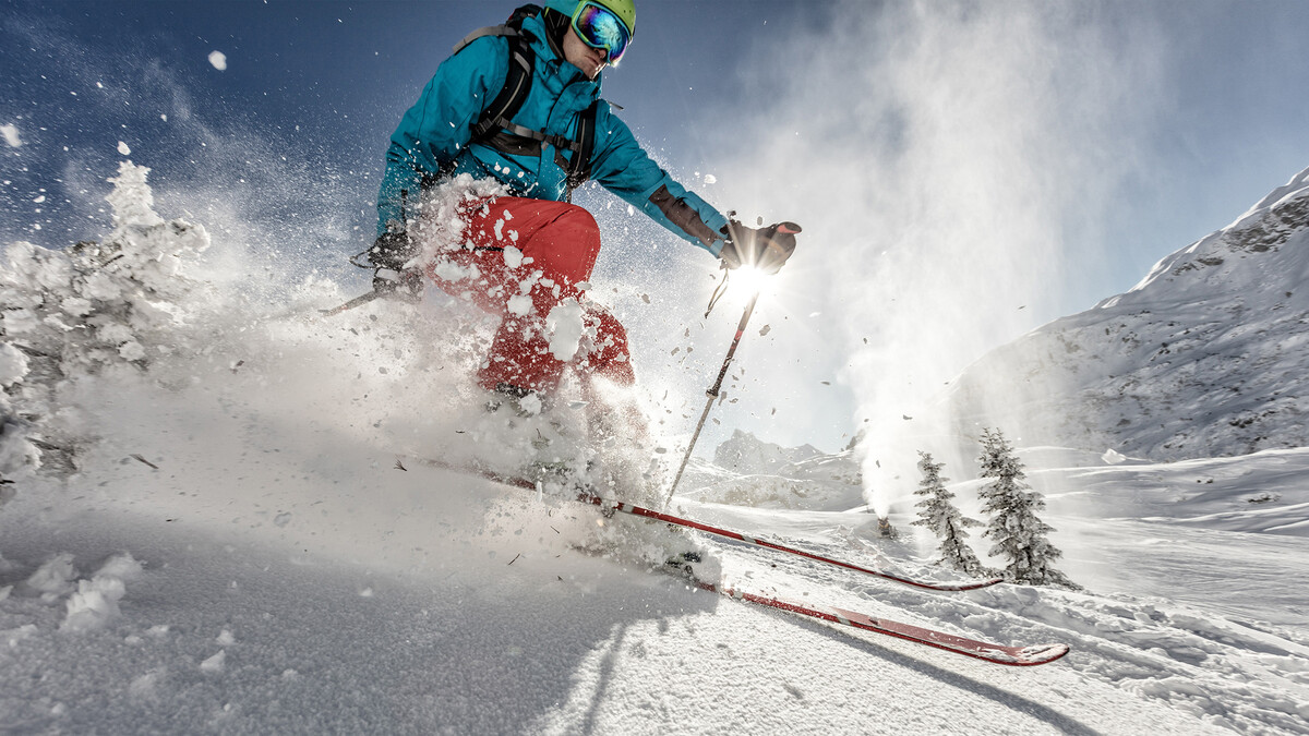 Ein Skifahrer fährt auf einen Skien durch den Tiefschnee, welcher entgegen spritzt. Hinter ihm sind Bäume und Berge zu sehen. Alles ist überdeckt mit Schnee. Diese Umgebung befindet sich in der Nähe vom Hotel Grand Resort Bad Ragaz, einem Superior Luxusresort in der Schweiz.