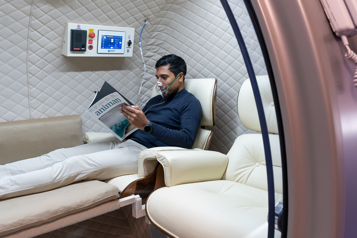 Im HBOT des Medizinischen Zentrums befindet sich ein Model. Er trägt eine Atemmaske, da er eine Sauerstofftherapie macht. Die Innenausstattung ist eher in weiss gehalten. Der Mann trägt eine weisse Hose und einen dunkelblauen Pullover. Das Medizinische Zentrum gehört zum Wellnesshotel Grand Resort Bad Ragaz, einem Superior Luxusresort in der Schweiz.