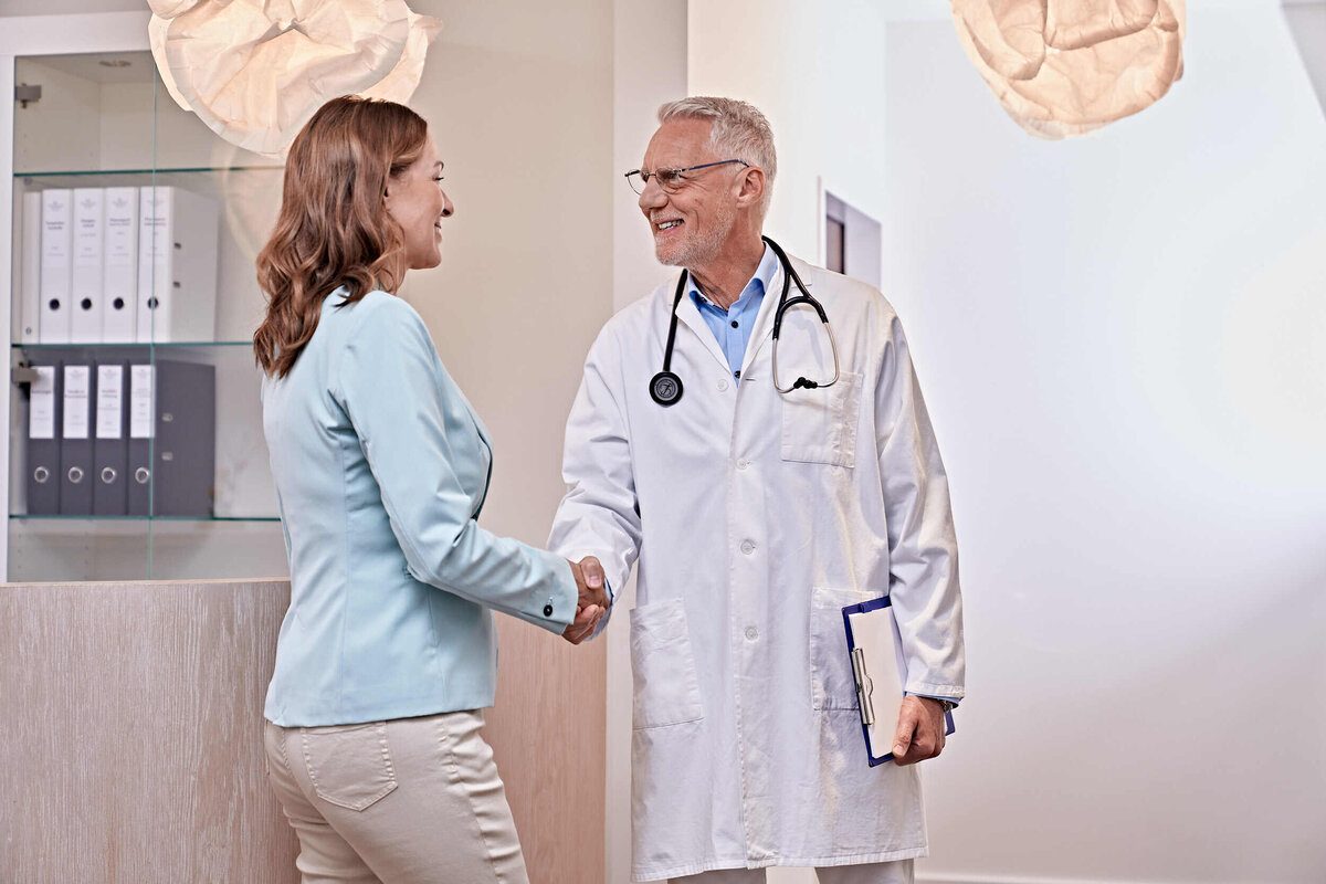 Ein Arzt in einem weißen Kittel, der ein Klemmbrett in der Hand hält, gibt einer Frau in einem Büro herzlich die Hand und vermittelt dabei ein Gefühl von Vertrauen und Professionalität.