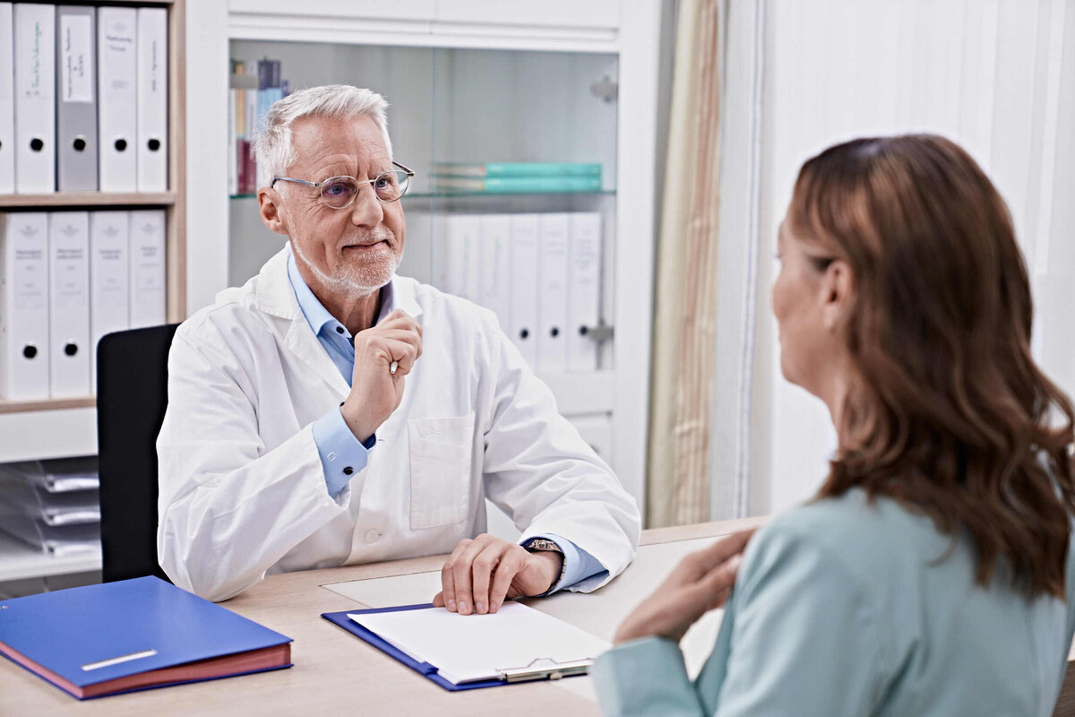 Ein Arzt berät eine Kundin an einem Tisch. Der Doktor hat ein Weisen Kittel an und darunter ein hellblaues Hemd. Die Kundin schaut den Arzt an und ist mit dem Rucken zur Kamera.