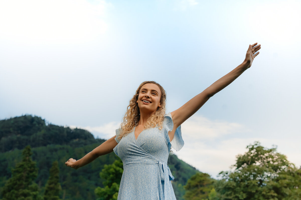 Eine Frau in einem hellblauen Kleid streckt freudig ihre Arme in einer üppigen Berglandschaft aus. Der Himmel ist strahlend blau und vermittelt ein Gefühl von Freiheit und Glück.