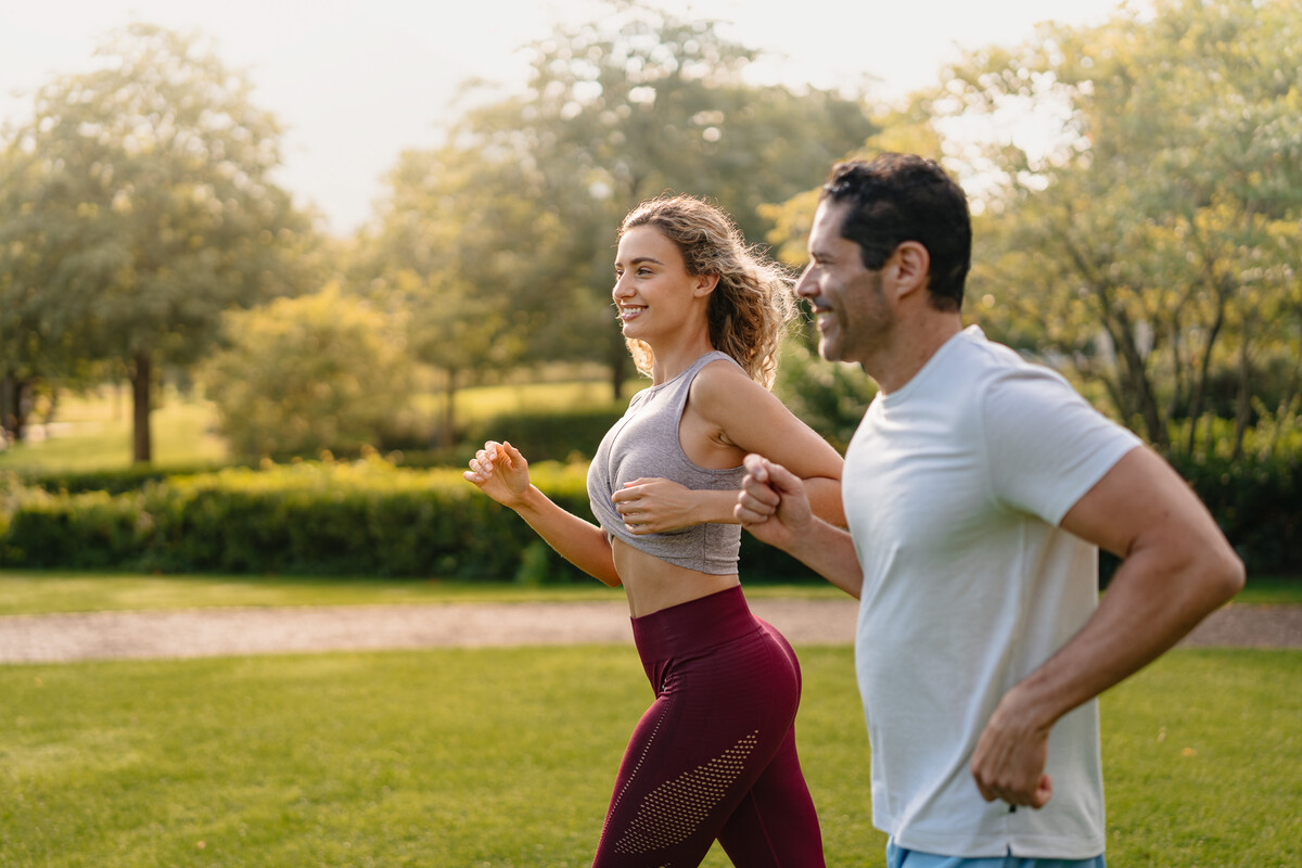 Eine Frau und ein Mann joggen an einem sonnigen Tag nebeneinander in einem Park, umgeben von üppigem Grün. Beide wirken fröhlich und voller Energie.