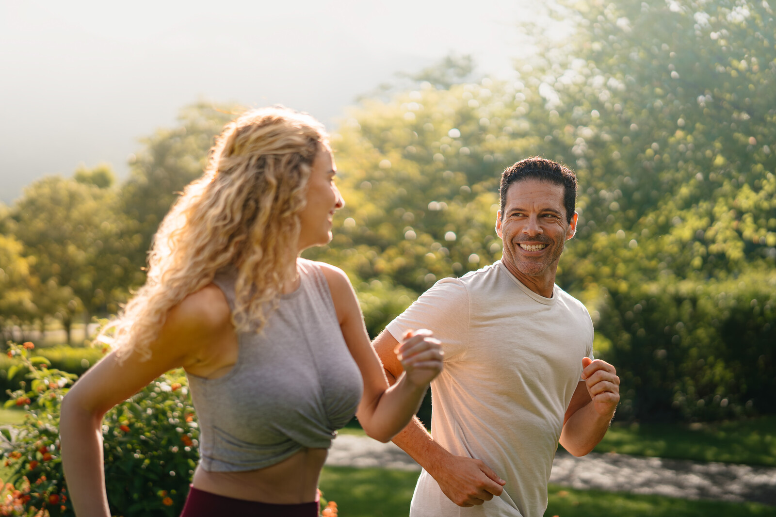 Ein Mann und eine Frau joggen draussen in einem sonnigen Park und lächeln sich an. Üppige Bäume und viel Grün schaffen eine lebendige, aufmunternde Atmosphäre.