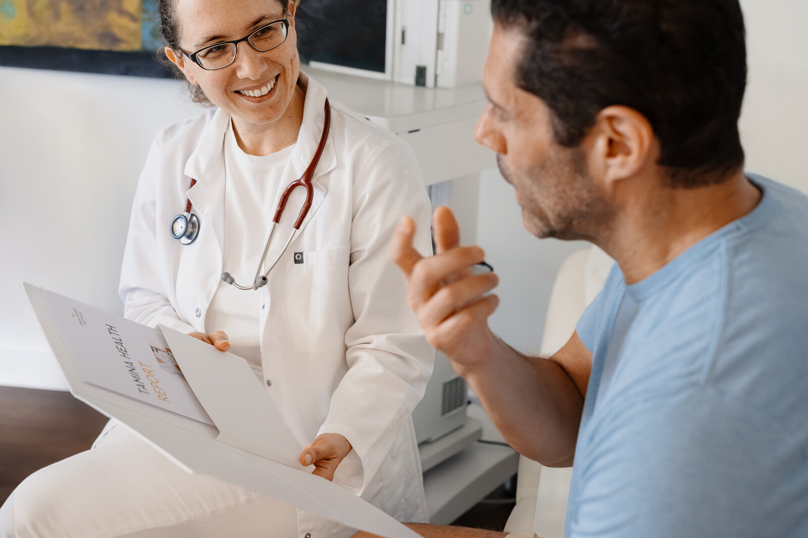 Ein Arzt in einem weissen Kittel mit einem Stethoskop lächelt, während er einem männlichen Patienten eine Broschüre zeigt. Sie scheinen ruhig über Gesundheit zu sprechen.