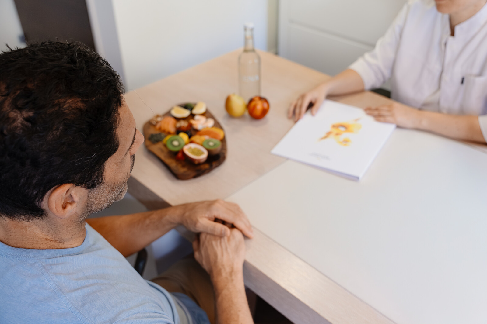Ein Mann und eine Ernährungsberaterin sitzen an einem Tisch. Auf einem Brett sind frische Früchte ausgestellt. Die Ernährungsberaterin hält ein Dokument mit einem Lebensmitteldiagramm in der Hand.