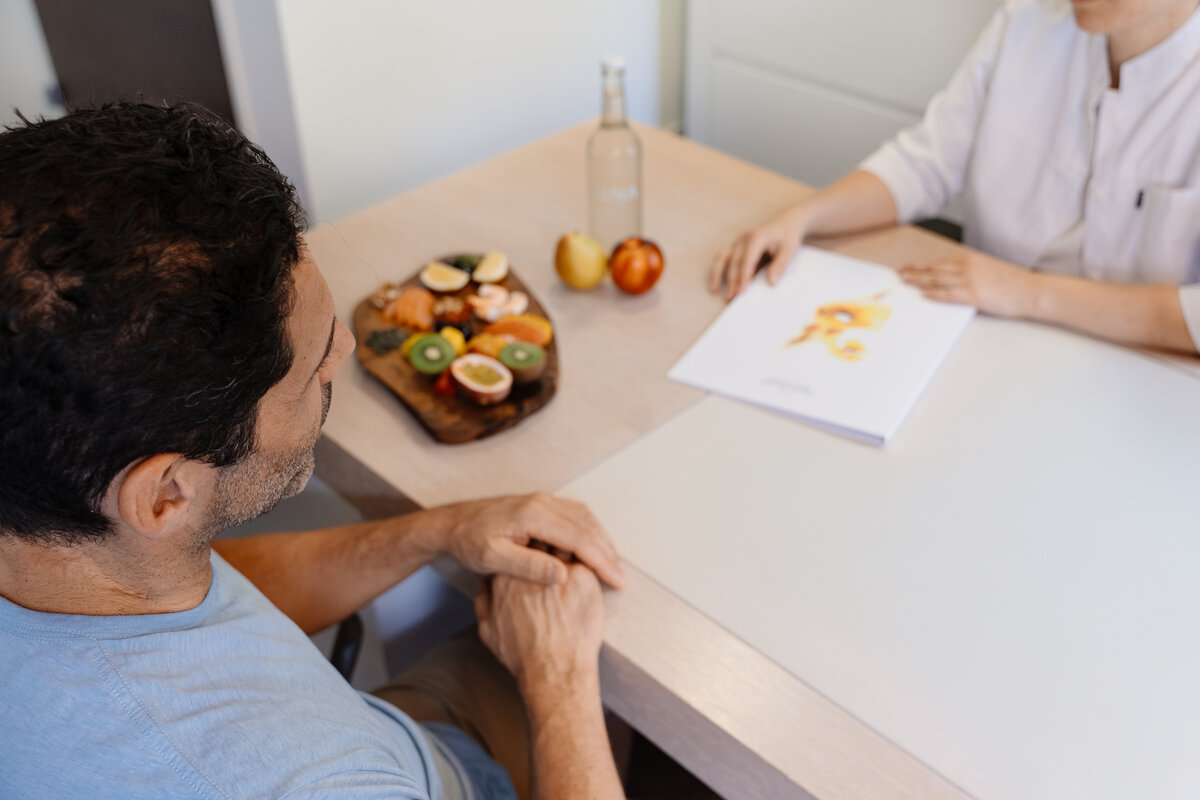 Ein Mann und eine Ernährungsberaterin sitzen an einem Tisch. Auf einem Brett sind frische Früchte ausgestellt. Die Ernährungsberaterin hält ein Dokument mit einem Lebensmitteldiagramm in der Hand.