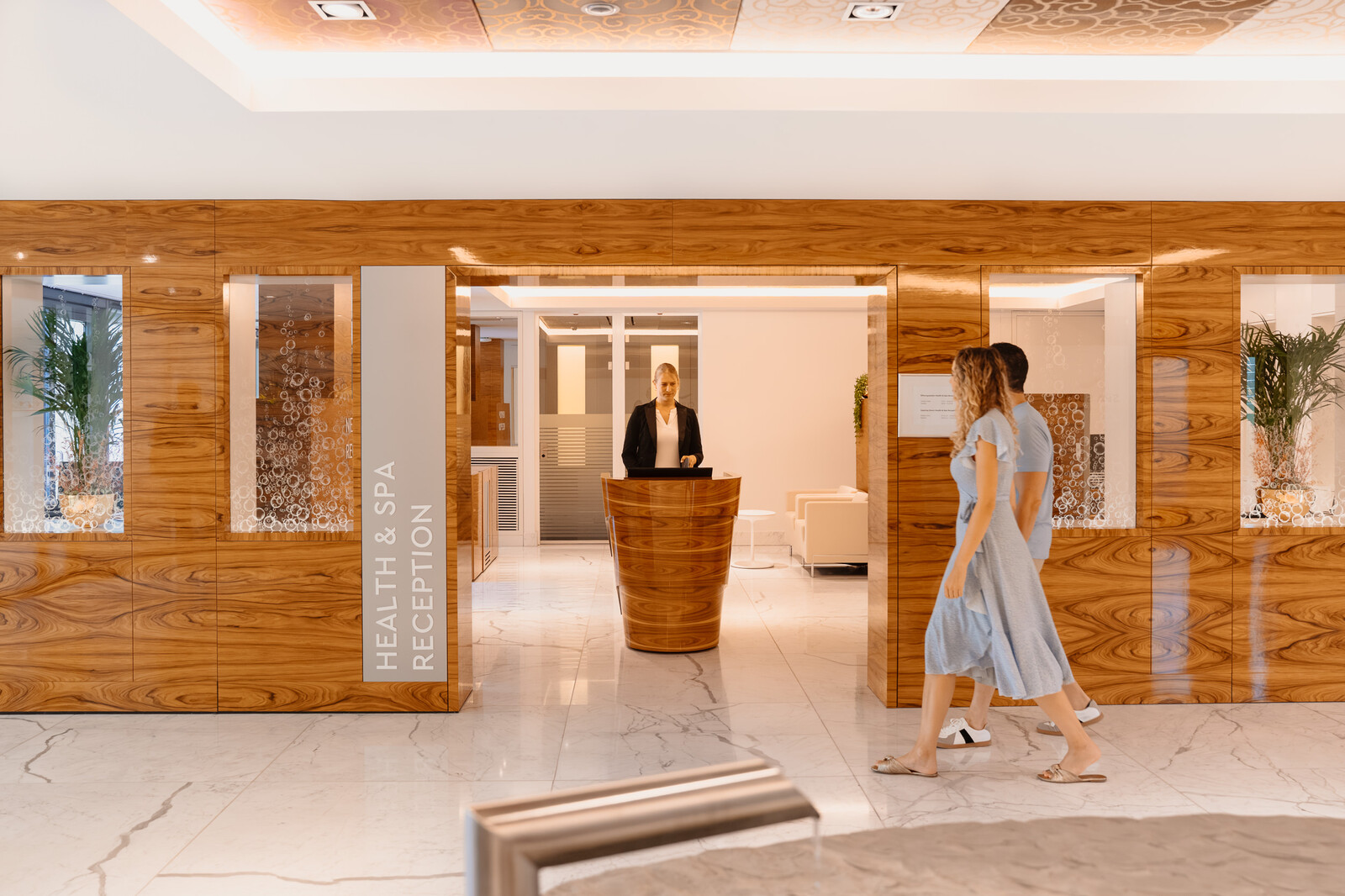 Der moderne Empfangsbereich der Health & Spa Reception für ein Gesundheits- und Wellnesszentrum mit Holzvertäfelung. Eine Empfangsdame steht hinter einem geschwungenen Schreibtisch, während zwei Personen auf einem Marmorboden vorbeigehen.