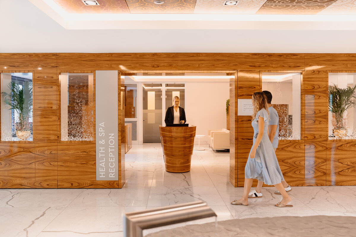 Der moderne Empfangsbereich der Health & Spa Reception für ein Gesundheits- und Wellnesszentrum mit Holzvertäfelung. Eine Empfangsdame steht hinter einem geschwungenen Schreibtisch, während zwei Personen auf einem Marmorboden vorbeigehen.